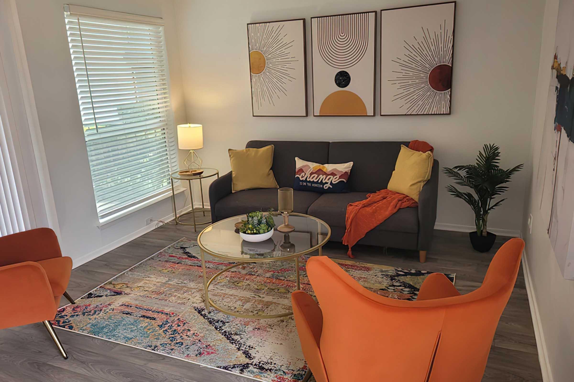 A cozy living room featuring a gray sofa with yellow pillows and a decorative throw. Two orange armchairs sit nearby, with a coffee table in front holding a small plant. Colorful artwork adorns the walls, and a lamp provides warm lighting, creating an inviting atmosphere. A patterned rug adds vibrancy to the space.