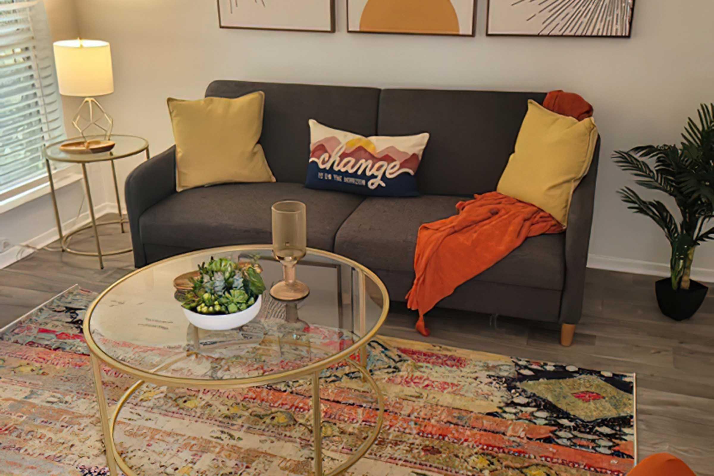 Cozy living room scene featuring a gray couch with yellow pillows, a decorative throw blanket in orange, and a glass coffee table. The table holds a small plant and a candle. A colorful area rug adds warmth, while sunlight pours in through nearby windows with sheer curtains. Stylish wall art is displayed above the couch.