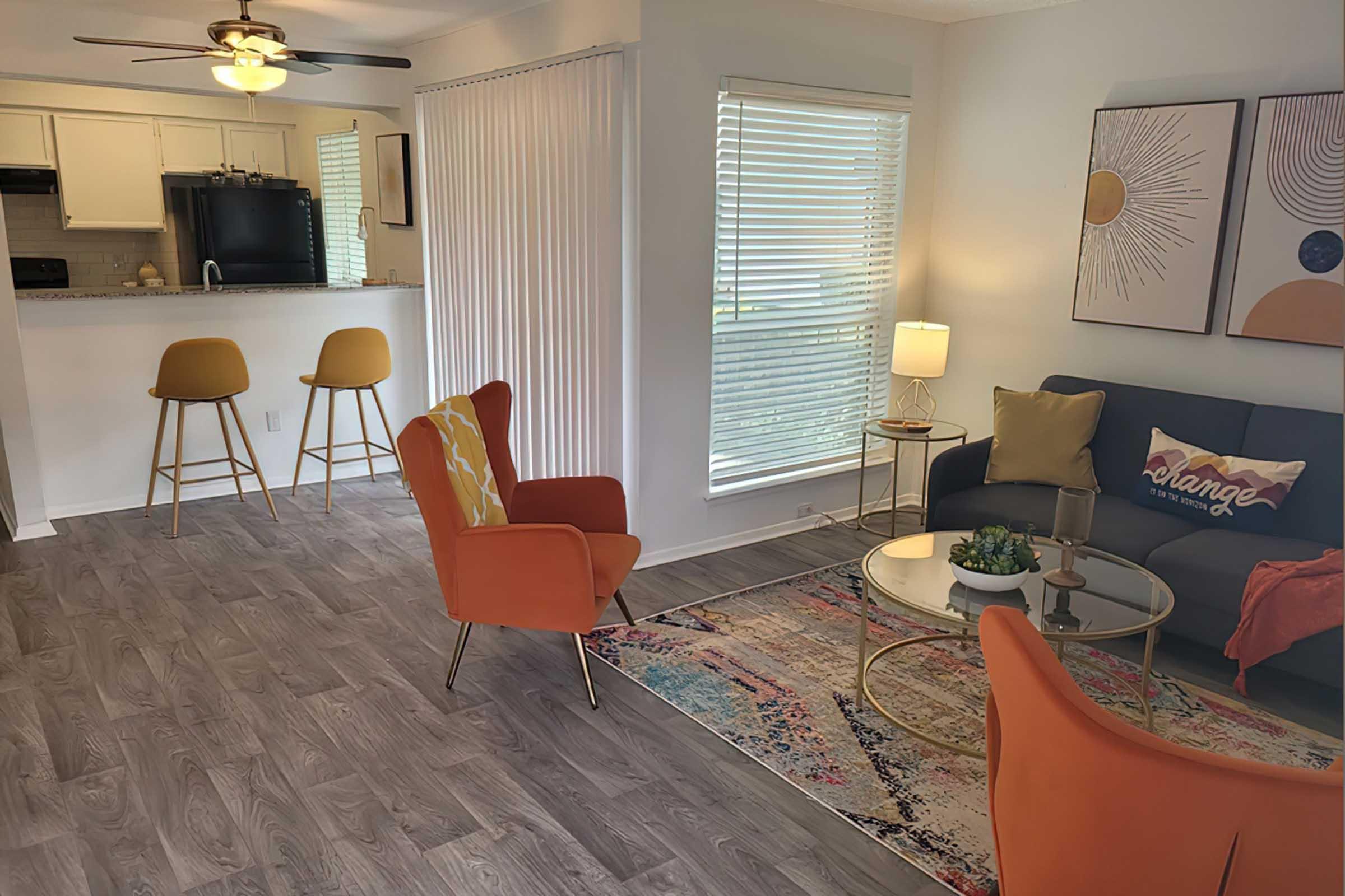 A stylish living room featuring two orange chairs and a navy sofa with decorative cushions. A round coffee table sits on a colorful rug, and there’s a standing lamp nearby. The background shows a kitchen area with wooden stools and a light color scheme, complemented by large windows allowing natural light.