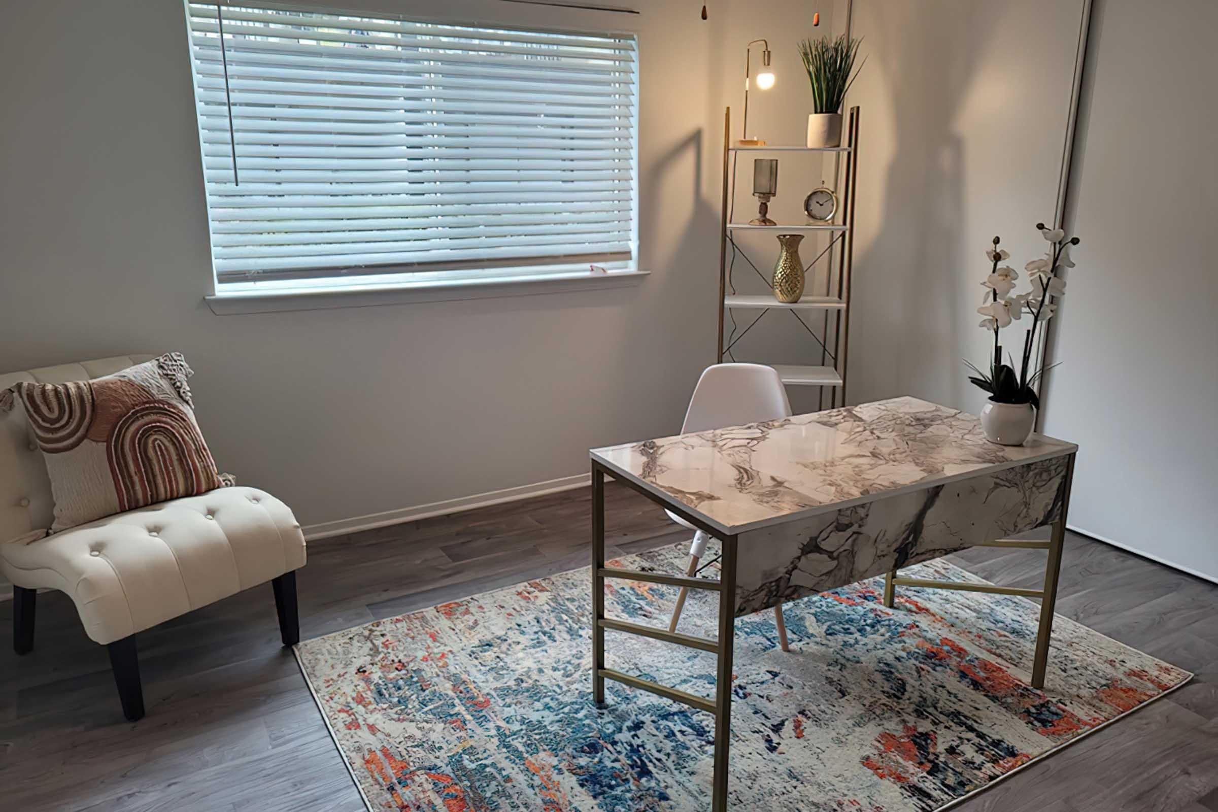 A modern home office featuring a marble-top desk with a small plant, a comfortable chair with decorative cushions, and a stylish shelving unit. The room is bright with natural light from a window covered by white blinds, and a colorful area rug adds warmth to the space.