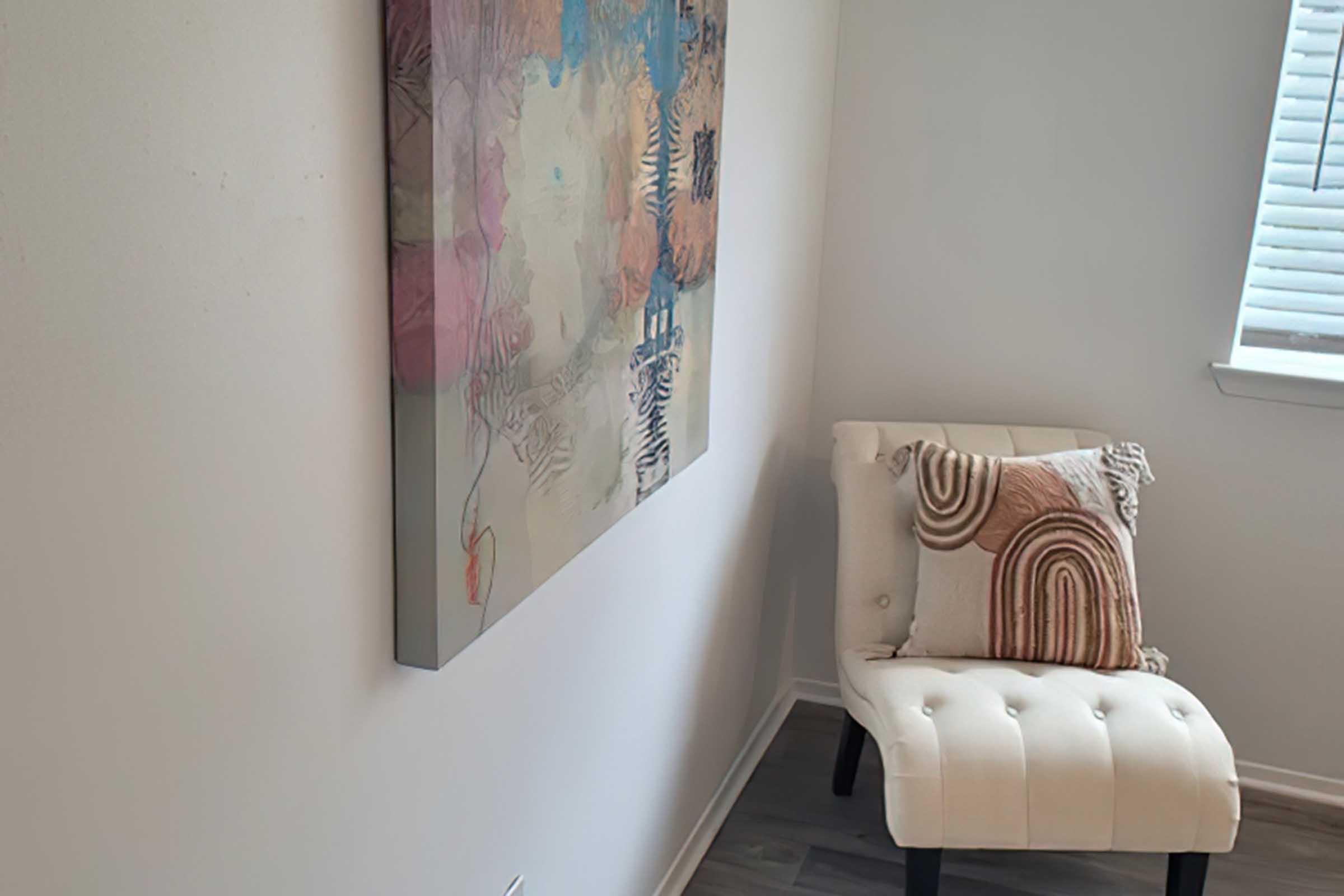 A cozy corner of a room featuring a light-colored chair with decorative pillows and an abstract painting on the wall. The painting includes soft colors and shapes, while the walls are a neutral tone. Natural light filters in through a window with blinds.