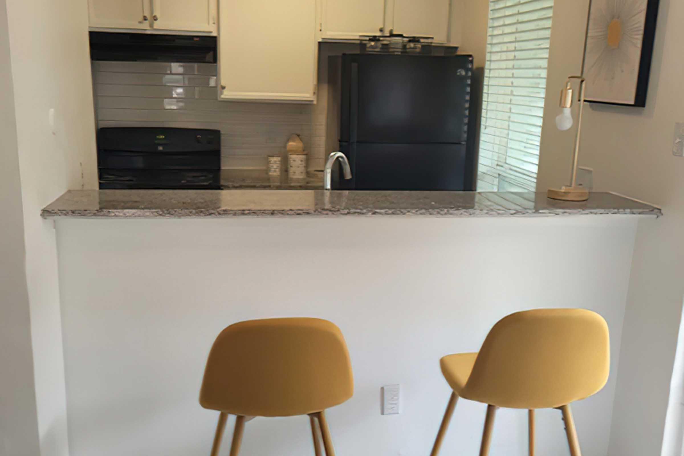 Modern kitchen with a granite countertop, featuring two yellow chairs. A black refrigerator and oven are visible in the background, along with light-colored cabinets and a window with blinds. A decorative lamp stands on the counter, adding a warm touch to the space.