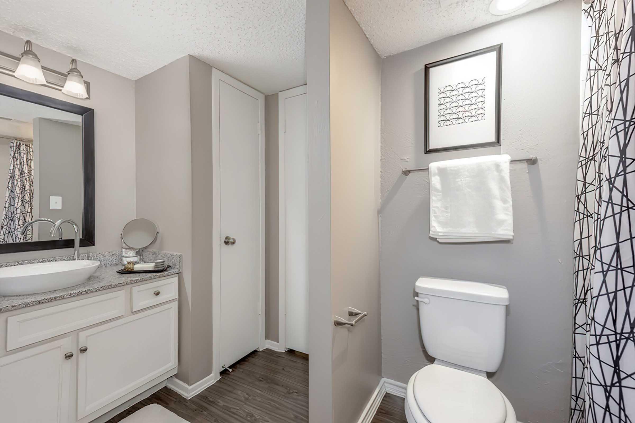 A modern bathroom featuring a double sink vanity with a mirror and countertop, light fixtures above, and a neatly organized space. On the right side, there is a toilet under a towel rack with a white towel. The walls are painted in neutral tones, enhancing the clean and contemporary look.