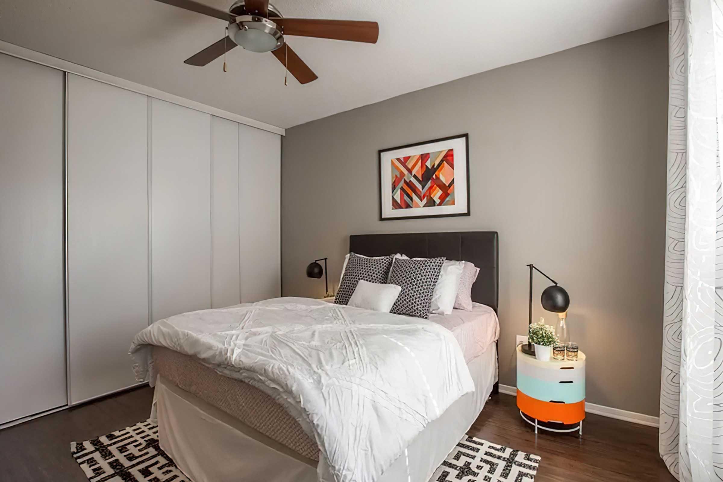 A cozy bedroom featuring a queen-sized bed with a light comforter and decorative pillows. There are nightstands on either side, each with a lamp. A colorful abstract artwork hangs on the wall above the bed. The room has a modern design with wood flooring and a patterned rug. Wardrobe doors are visible in the background.