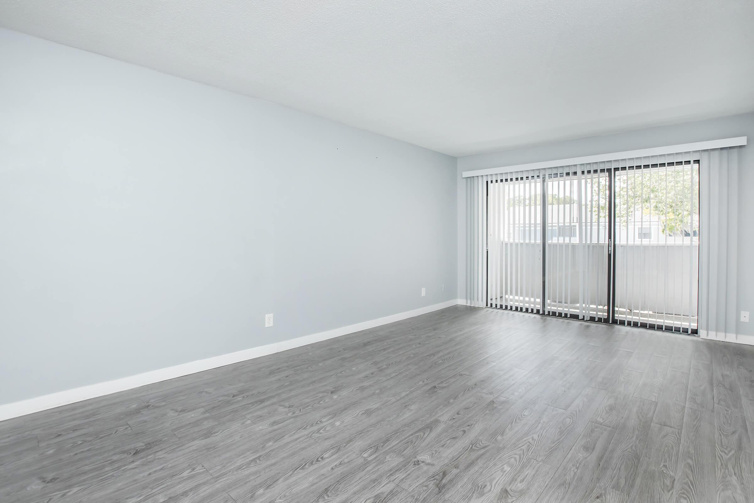 Spacious, empty living room with light gray walls and sleek gray flooring. Wide sliding glass doors with vertical blinds lead to a balcony, allowing natural light to fill the space. The room is uncluttered, offering a blank canvas for interior design.