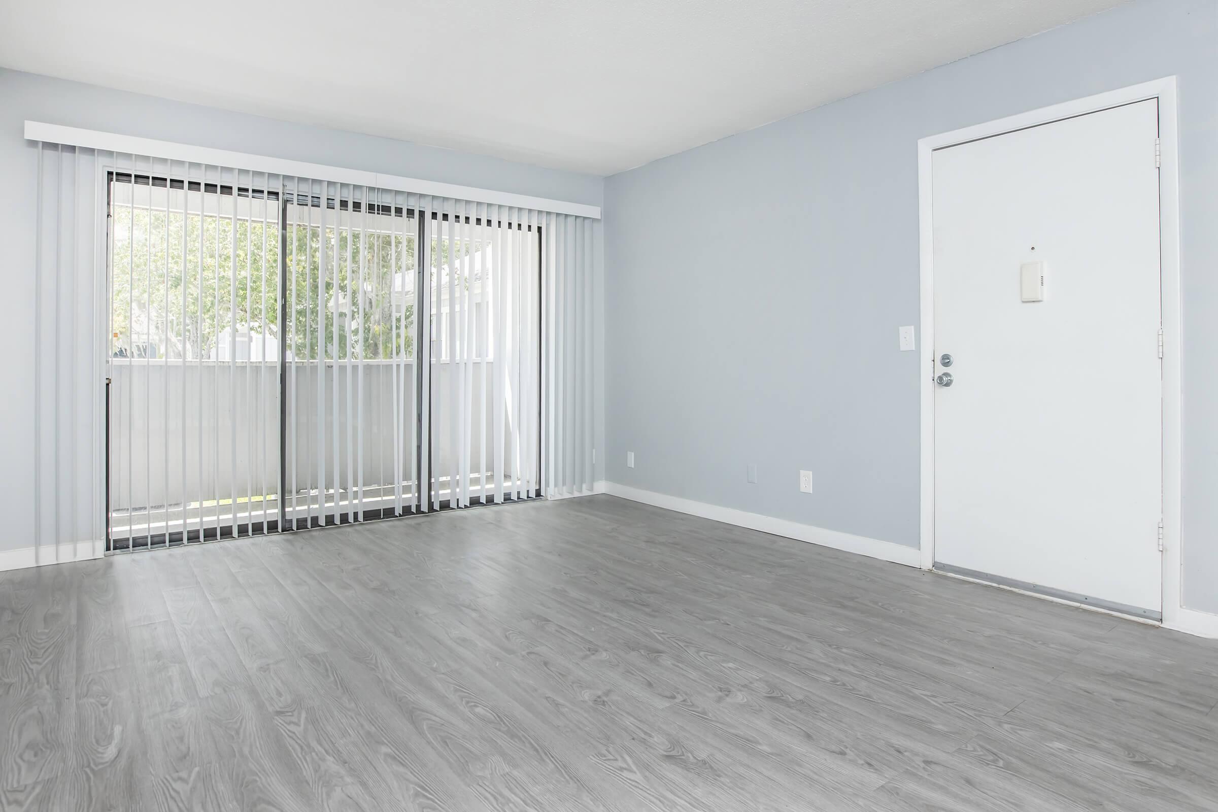 An empty room with light gray walls and a wooden floor. There is a large sliding glass door with vertical blinds leading to a balcony, and a plain white door on the right. The space is well-lit with natural light coming in from the balcony.