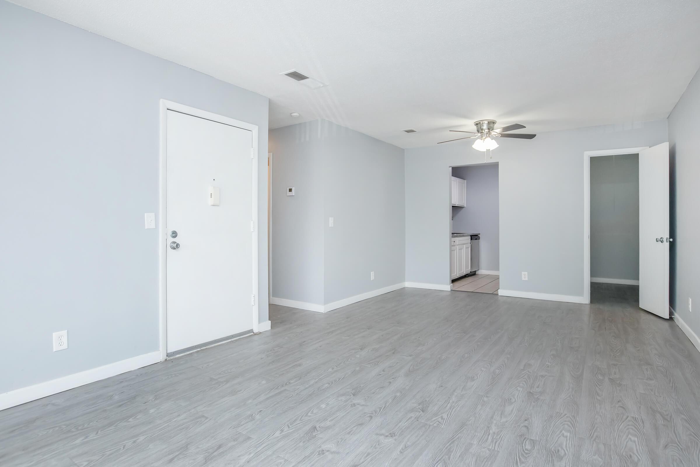 A spacious, empty room with light gray walls and flooring. A white door with a round handle is on the left, and an open doorway leads to another area. A ceiling fan is mounted above, and a kitchen can be seen in the background through the open door. The space is well-lit and uncluttered.