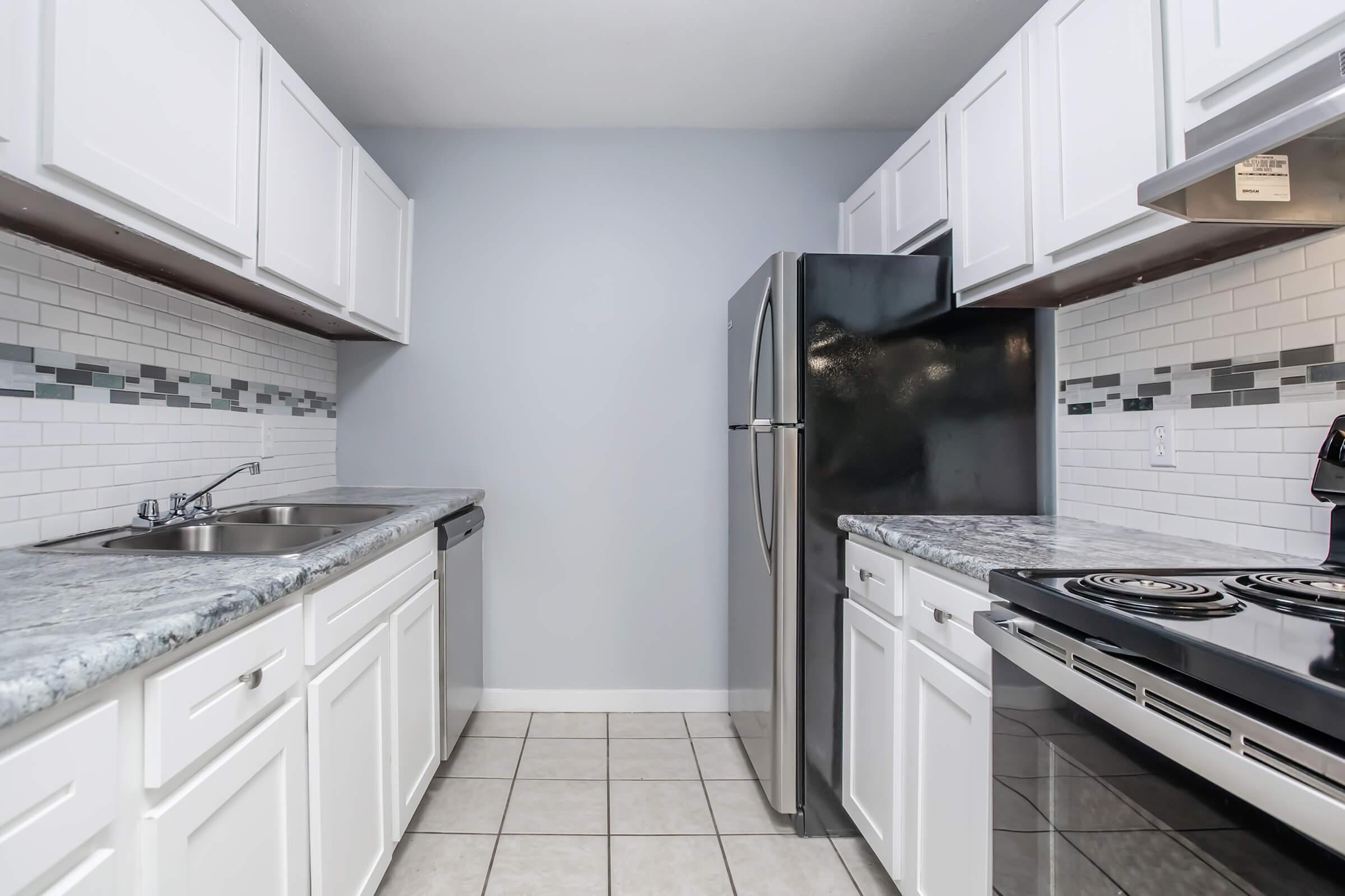 A modern kitchen featuring white cabinetry, gray walls, and a stainless steel refrigerator. The countertops are speckled gray, and the backsplash includes a mix of white and gray tiles. The kitchen includes a sink, stove, and ample cabinet space, with tiled flooring.