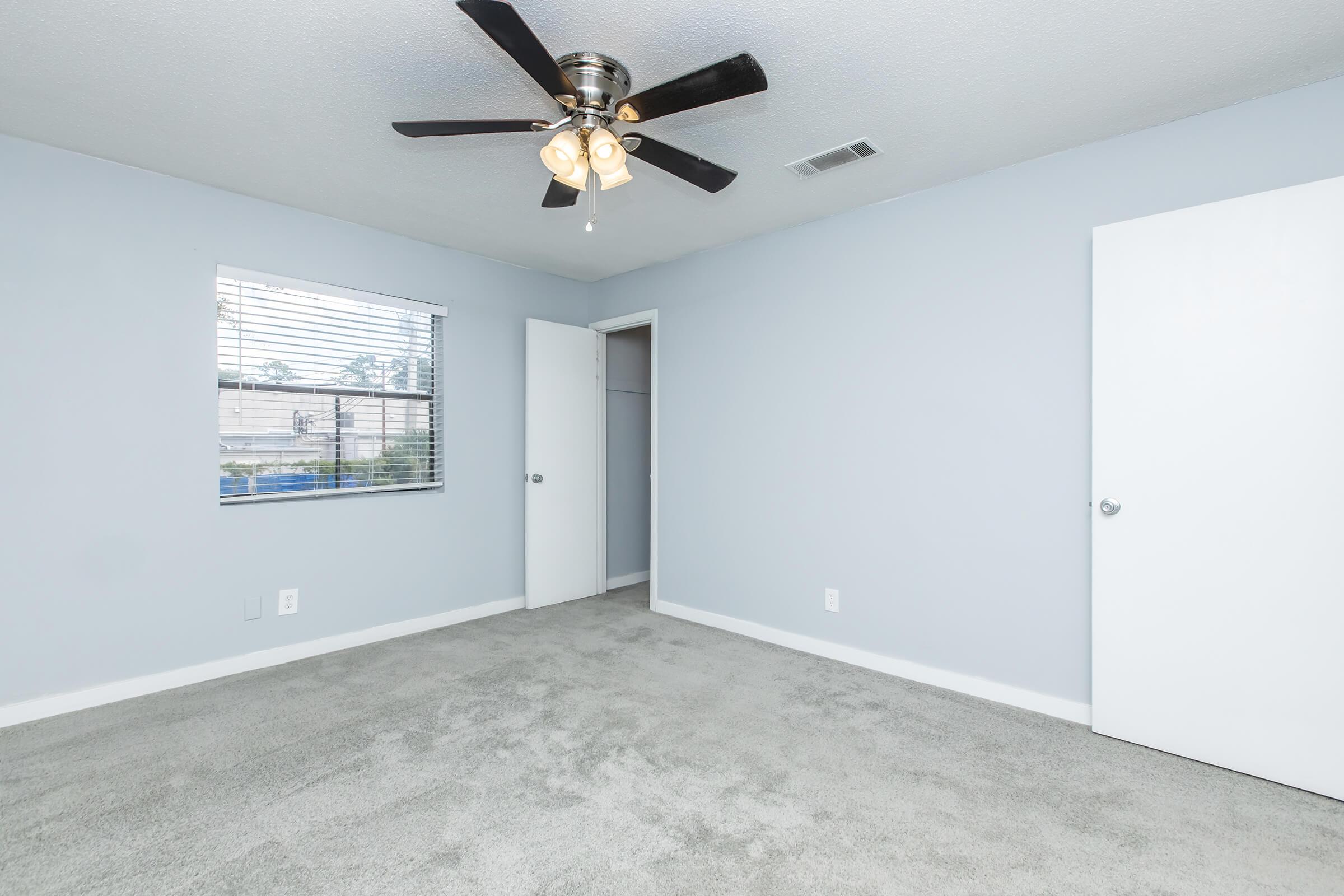 A spacious, light blue bedroom featuring a ceiling fan with light fixtures, gray carpeting, and a large window with horizontal blinds. There are two doors: one leading to a closet and the other to the room's exit. The walls are painted a soft blue, creating a calm and inviting atmosphere.