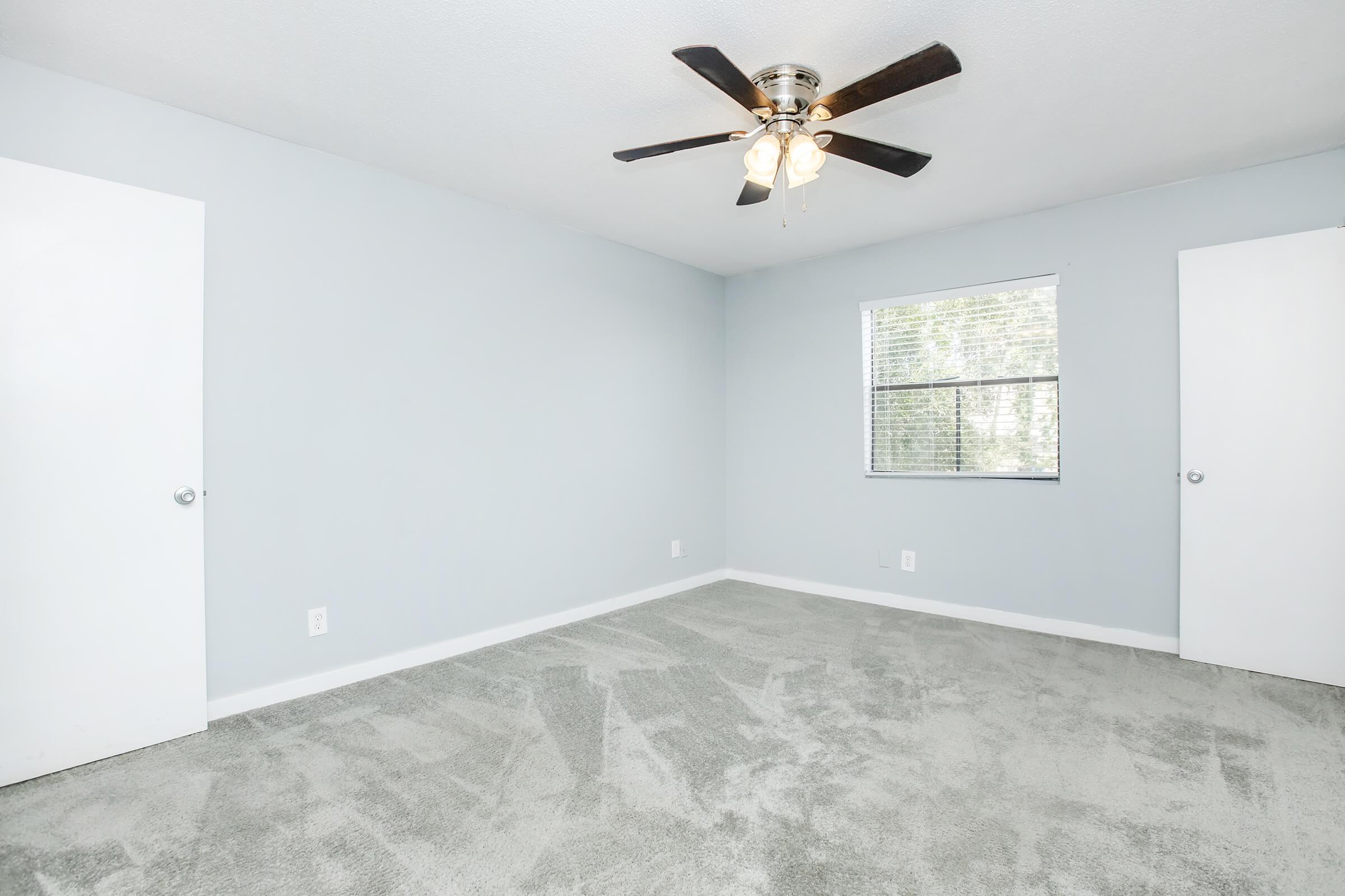 Empty bedroom featuring light gray walls, plush carpet, and a ceiling fan with light. Two white doors are visible on either side, and a window provides natural light, showcasing a view of greenery outside. The room appears tidy and ready for decoration or furnishing.