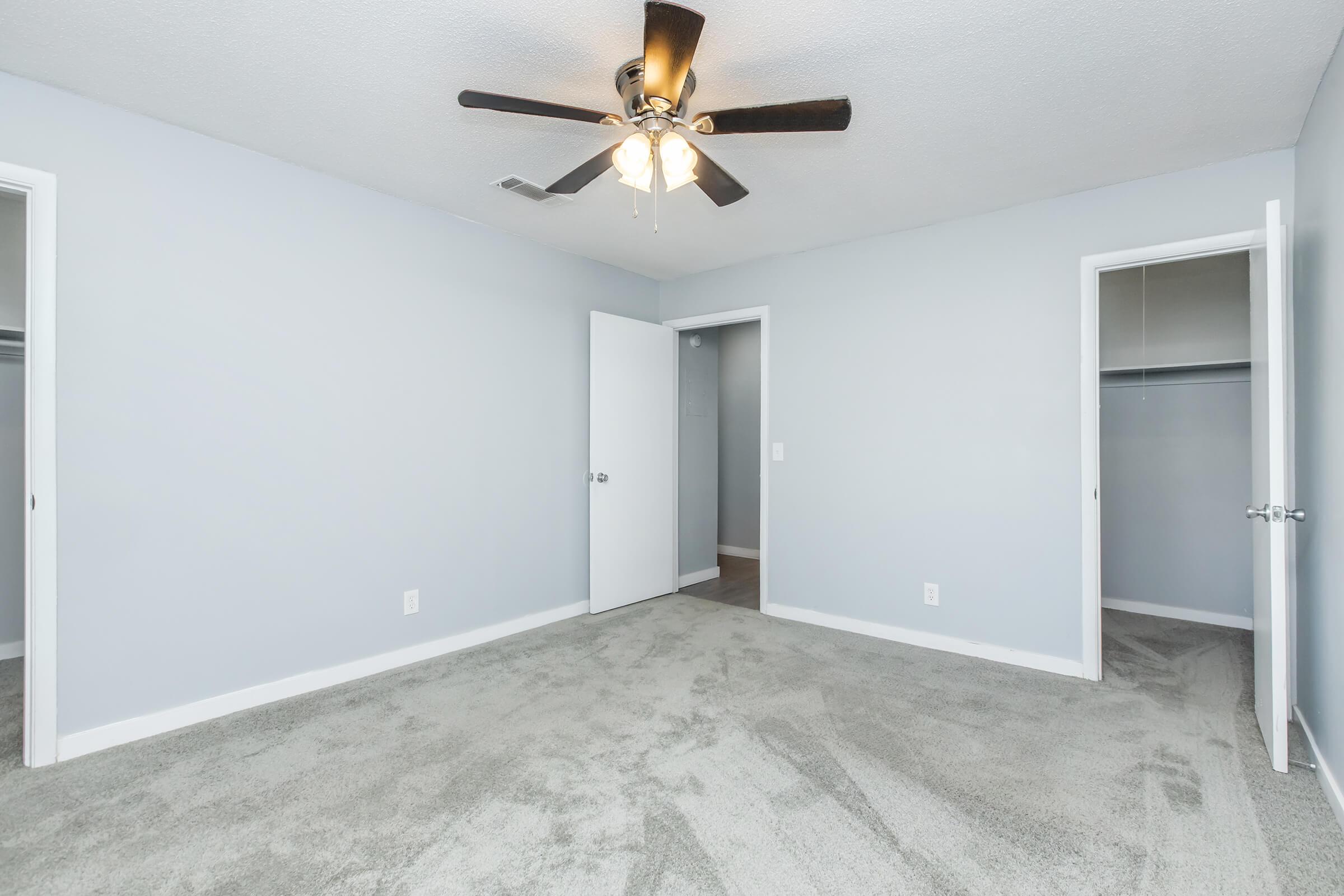 A well-lit, empty bedroom featuring light gray walls and plush carpet. There is a ceiling fan with light, a closed door on one side, and an open closet space visible on the opposite side. The room is spacious with a clean, modern appearance, perfect for personalizing.