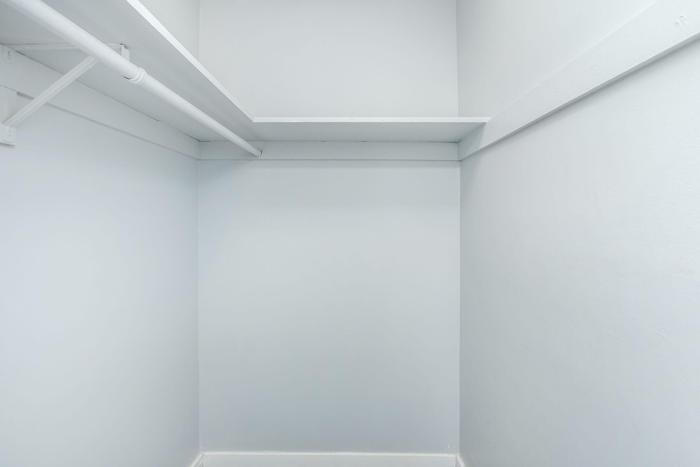 Empty closet with white walls and a simple shelf on the upper left side. The floor is bare and the space is illuminated with soft lighting, giving a clean and minimalist appearance.