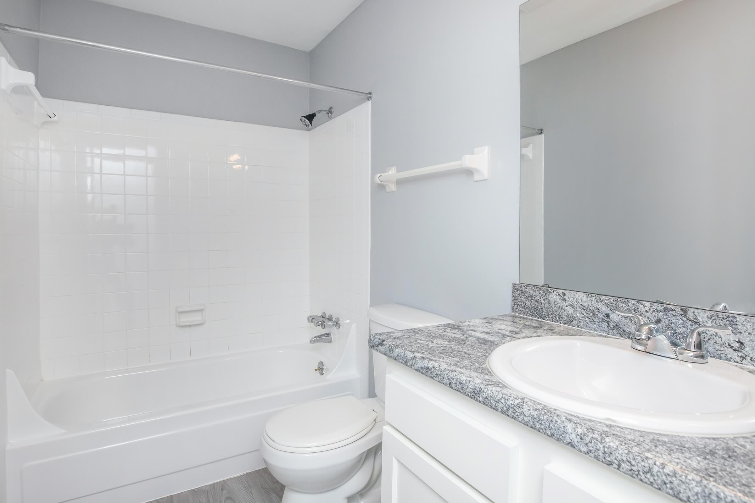 A clean and modern bathroom featuring a white tiled shower with a glass shower curtain, a granite countertop with a sink, and a white toilet. The walls are painted light gray, enhancing the spacious feel, and there's a mirror reflecting the room. Bright lighting adds to the fresh and inviting atmosphere.