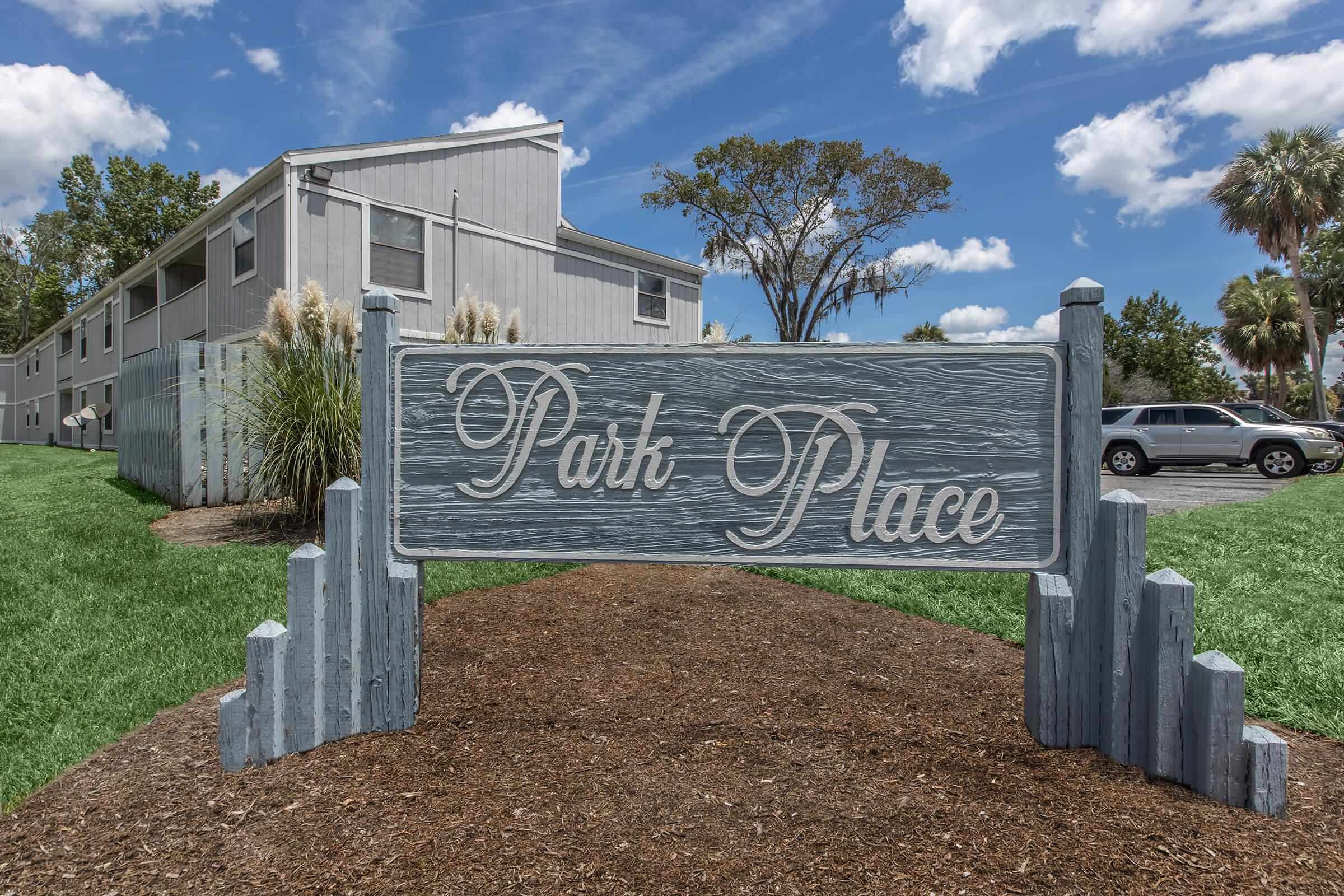 A sign reading "Park Place" stands in front of a landscaped area, featuring low shrubs and decorative stones. In the background, a multi-story building is visible under a blue sky adorned with clouds. The scene conveys a pleasant, well-maintained environment.