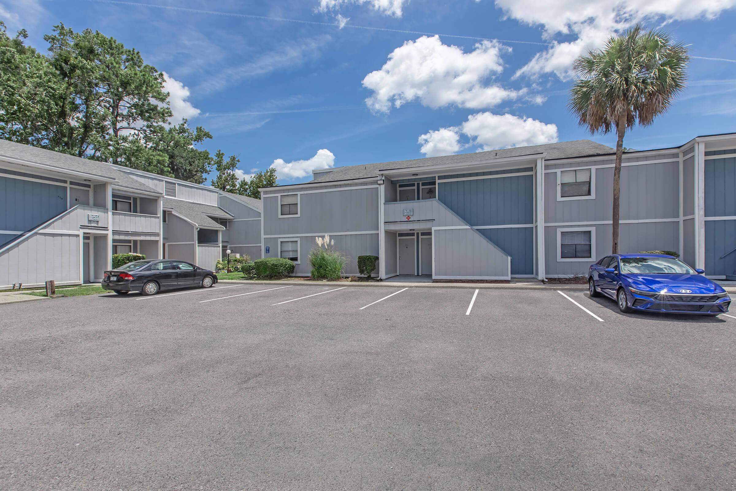 A view of a residential apartment complex with two buildings in light blue and gray colors. A parking lot in the foreground has three parked cars, and there are trees in the background under a partly cloudy sky.