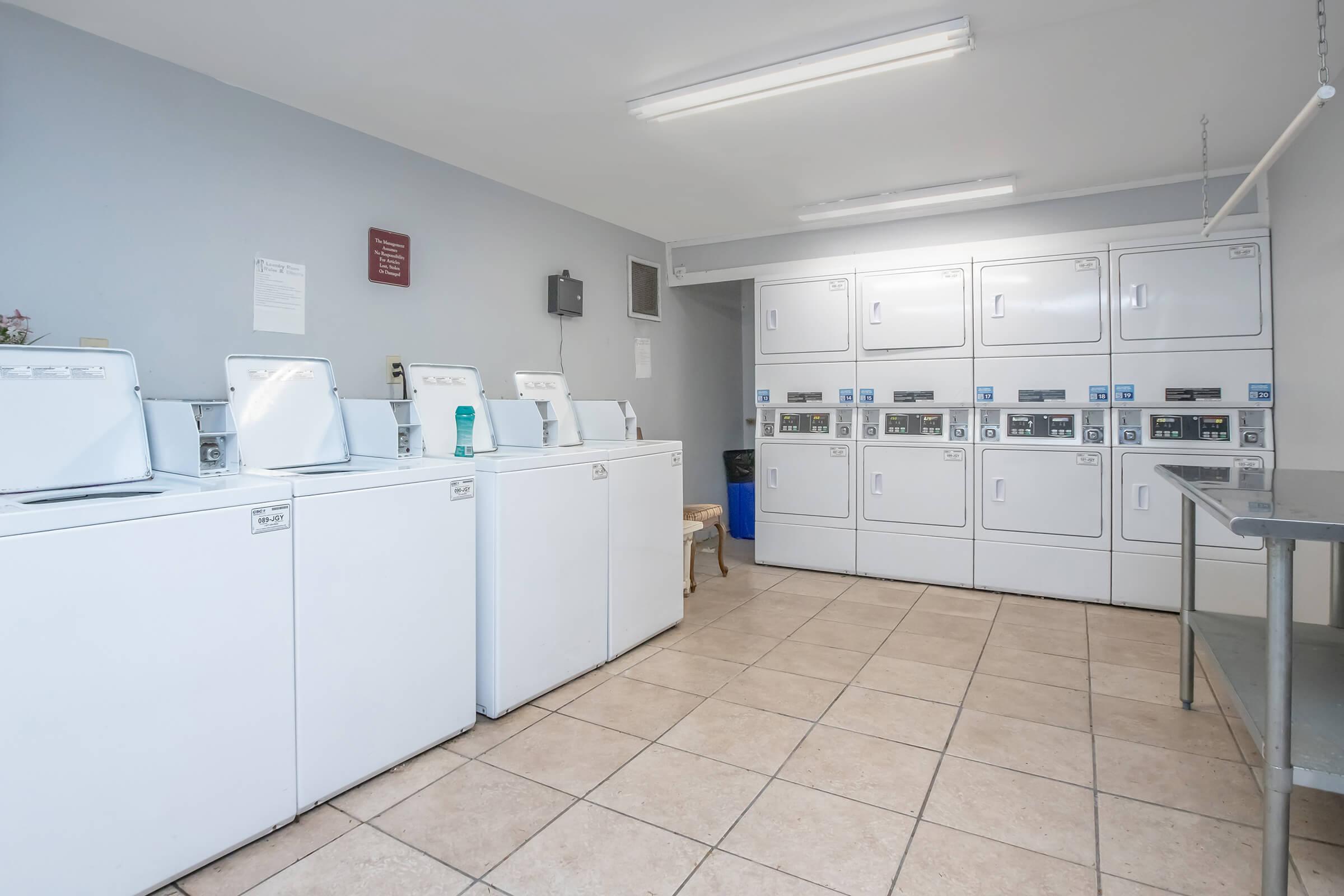A clean and well-lit laundry room featuring several white washing machines and dryers lined up against the wall. The floor is tiled, and there is a folding table on the side. There are notices on the walls and a chair in the corner, creating a functional space for doing laundry.
