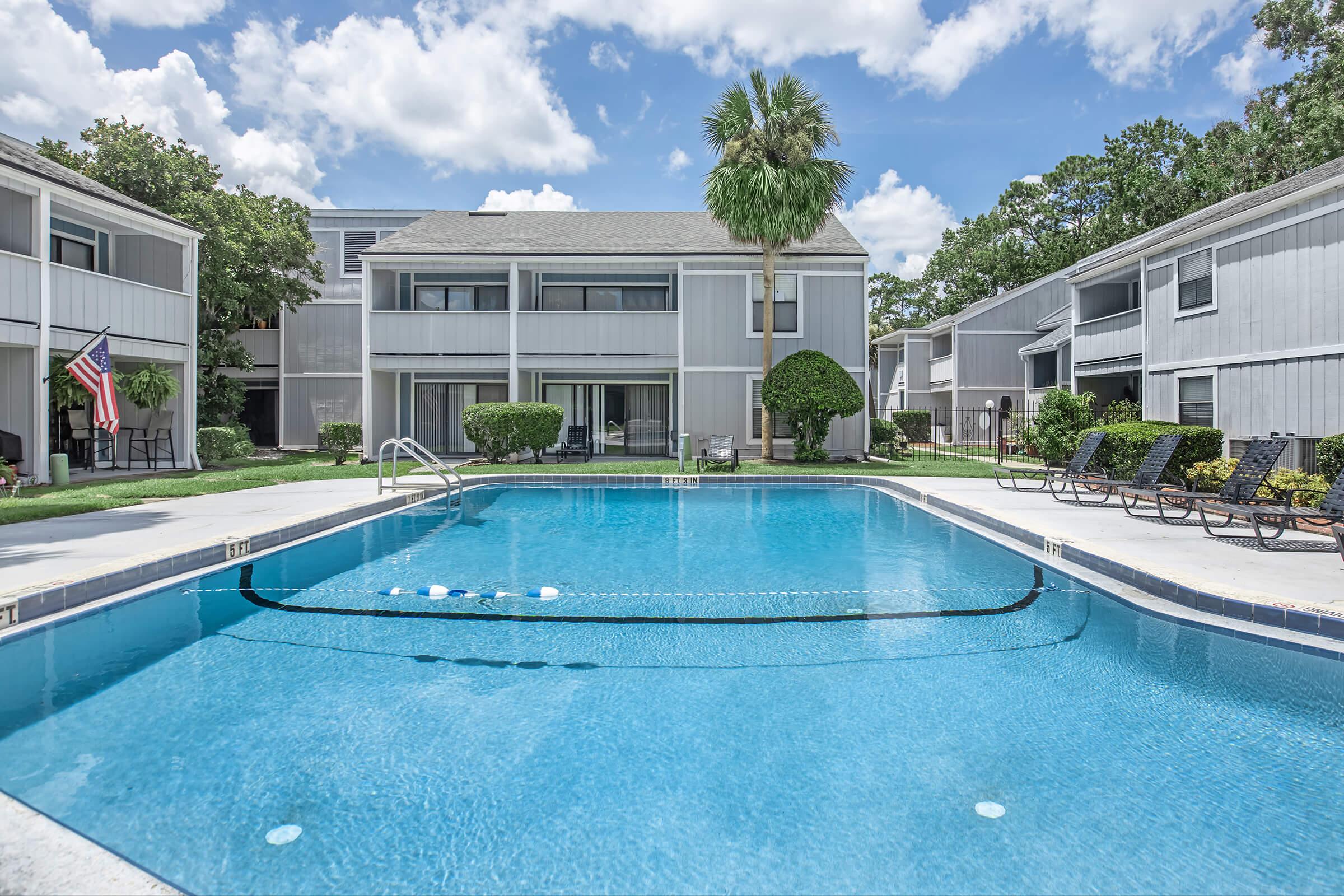 A sparkling blue swimming pool surrounded by lounge chairs, with a backdrop of well-maintained gray buildings and lush greenery. The sky is partly cloudy, adding a vibrant ambiance to the scene. An American flag can be seen in the background, enhancing the inviting atmosphere of the outdoor space.