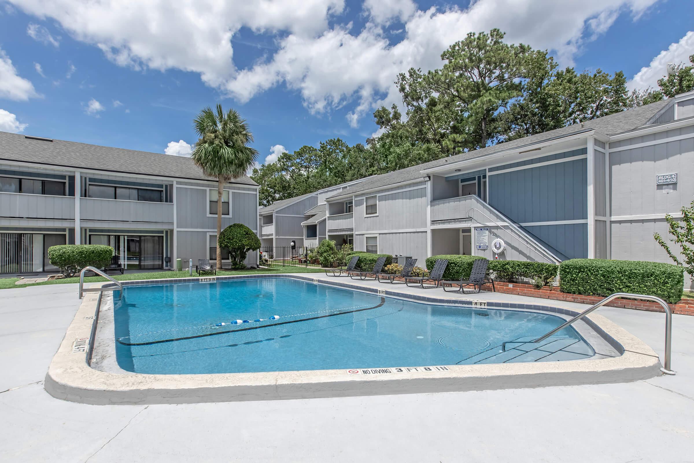 A clear blue swimming pool surrounded by lounge chairs in a landscaped area, with two multi-level residential buildings visible in the background and lush greenery and clouds in the sky above. The pool area is well-maintained and inviting.