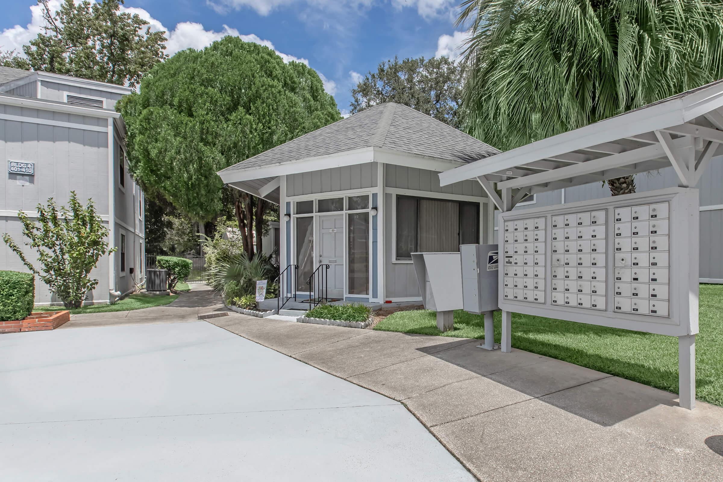 A well-maintained apartment complex entrance featuring a gray building with a welcoming porch. In the foreground, there is a set of mailboxes and a paved pathway leading to the entrance. Lush green trees and well-manicured landscaping enhance the inviting atmosphere under a bright blue sky.
