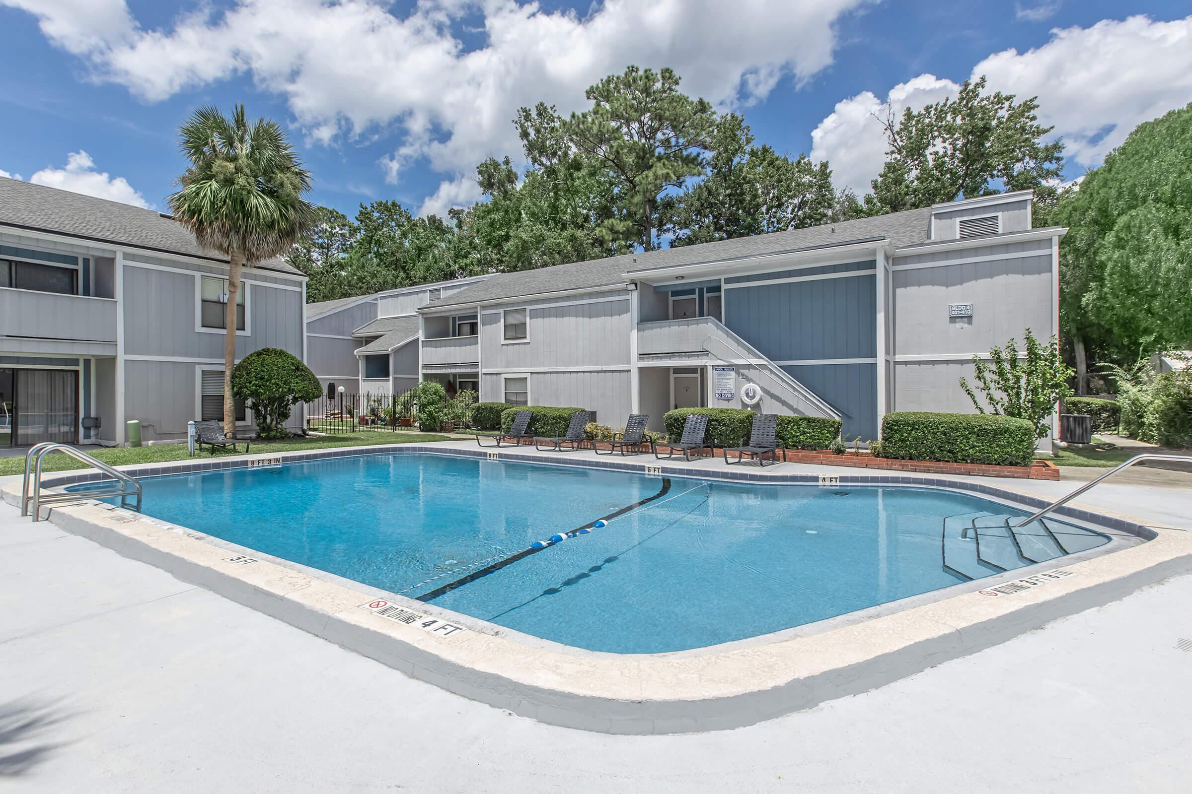 A swimming pool surrounded by lounge chairs in a residential area. The pool is clear and inviting, with lush greenery and trees in the background, under a bright blue sky with scattered clouds. Two buildings are visible, showcasing a tranquil apartment complex setting.