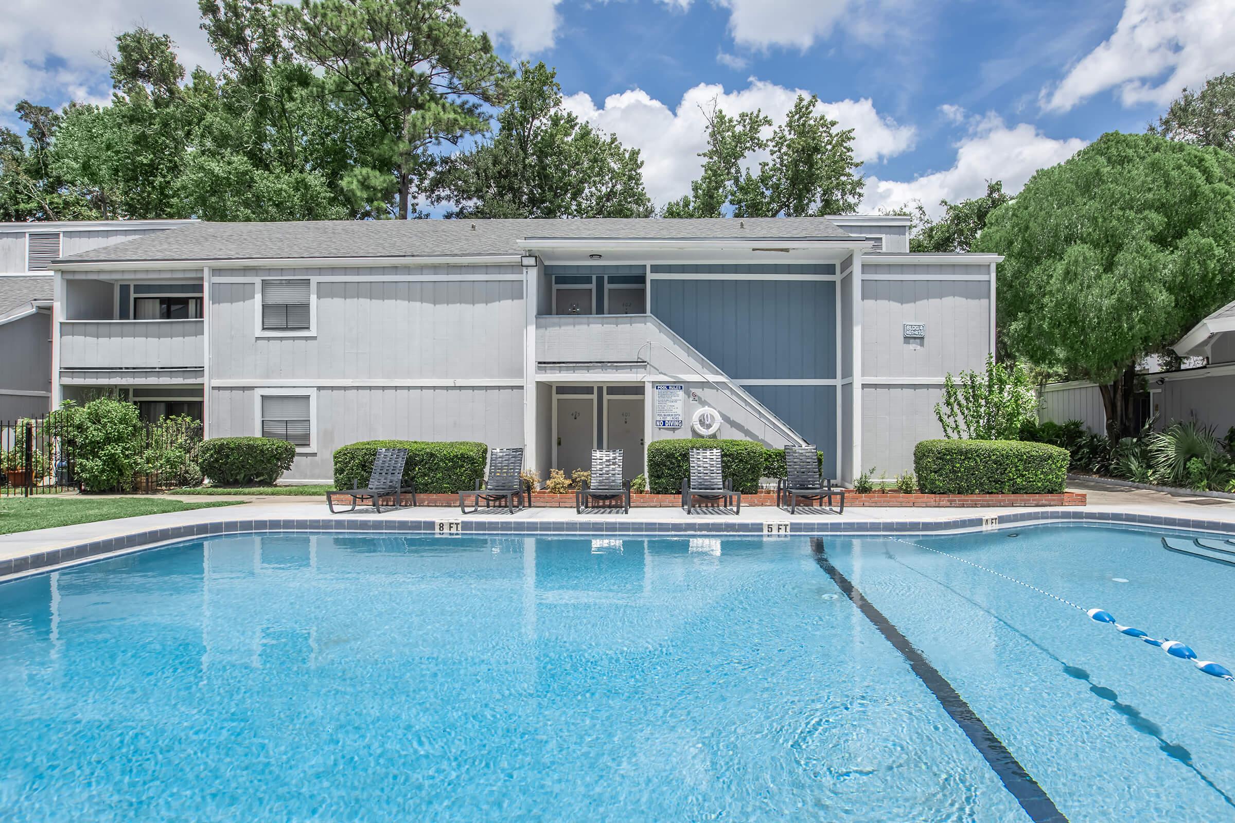 A view of an apartment complex with a swimming pool in the foreground. The building features several balconies, surrounded by well-maintained landscaping with bushes and trees. The sky is partly cloudy, reflecting a sunny day. Lounge chairs are positioned around the pool area.