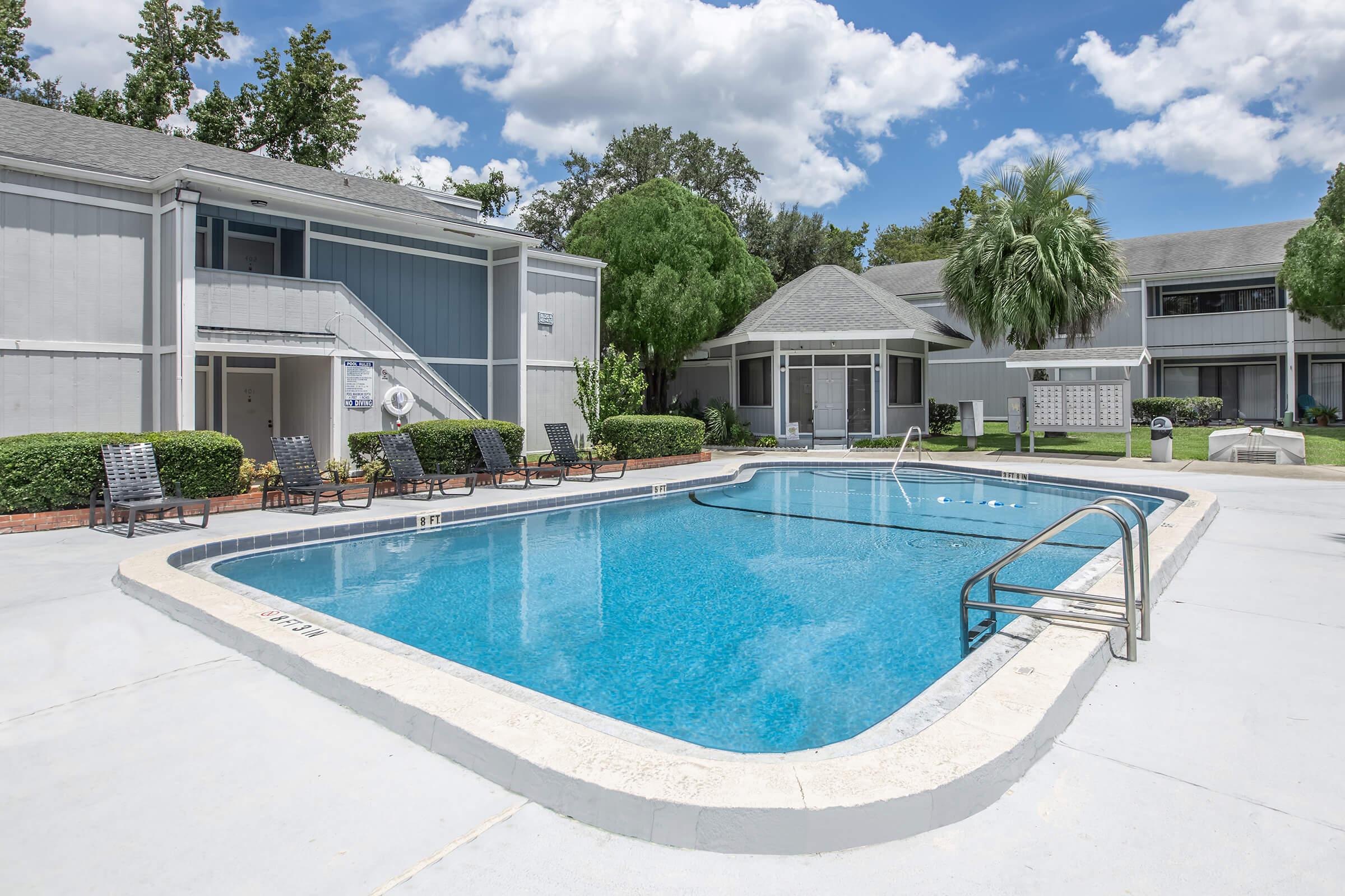 A clear blue swimming pool surrounded by a concrete deck, with lounge chairs on one side. In the background, there are gray multi-unit buildings with green landscaping, including palm trees and bushes. The sky is bright with scattered clouds.