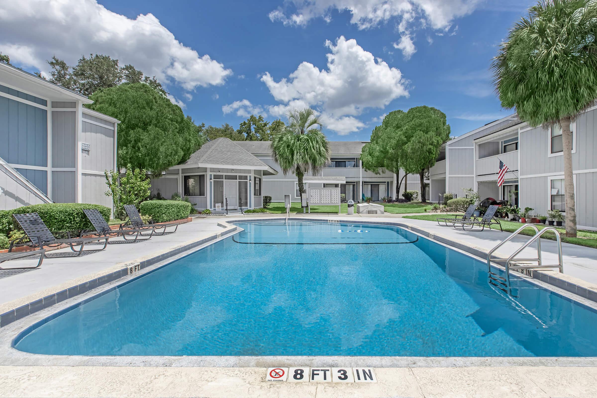 Swimming pool surrounded by lounge chairs and palm trees, with a clear blue sky above. The pool has a depth marker indicating 8 feet 3 inches. Nearby buildings are visible, and there are well-maintained green lawns. The scene conveys a relaxing outdoor setting.
