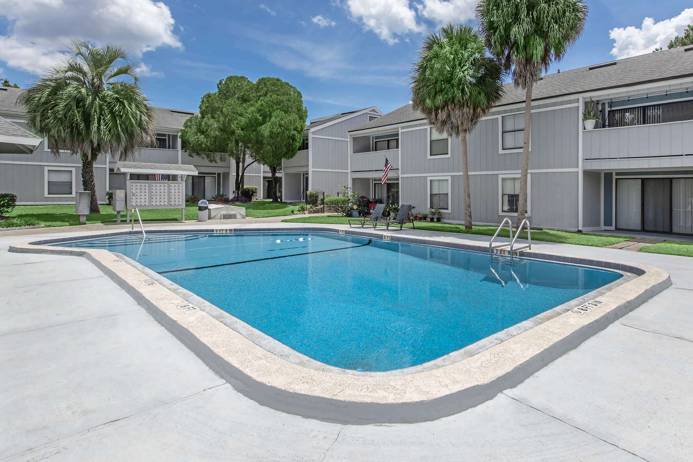A clear blue swimming pool surrounded by a concrete deck and landscaped greenery. In the background, there are gray apartment buildings with balconies and palm trees. The sky is bright with a few fluffy clouds.