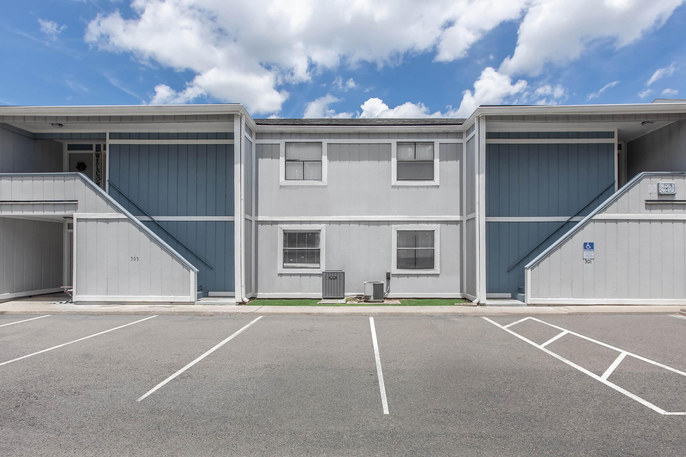 Two-story apartment building with blue and gray siding. The facade features two staircases leading to entrances on the upper level. A small air conditioning unit is visible on the ground level, and the area in front has designated parking spaces and patches of grass. Fluffy white clouds are scattered in a blue sky above.