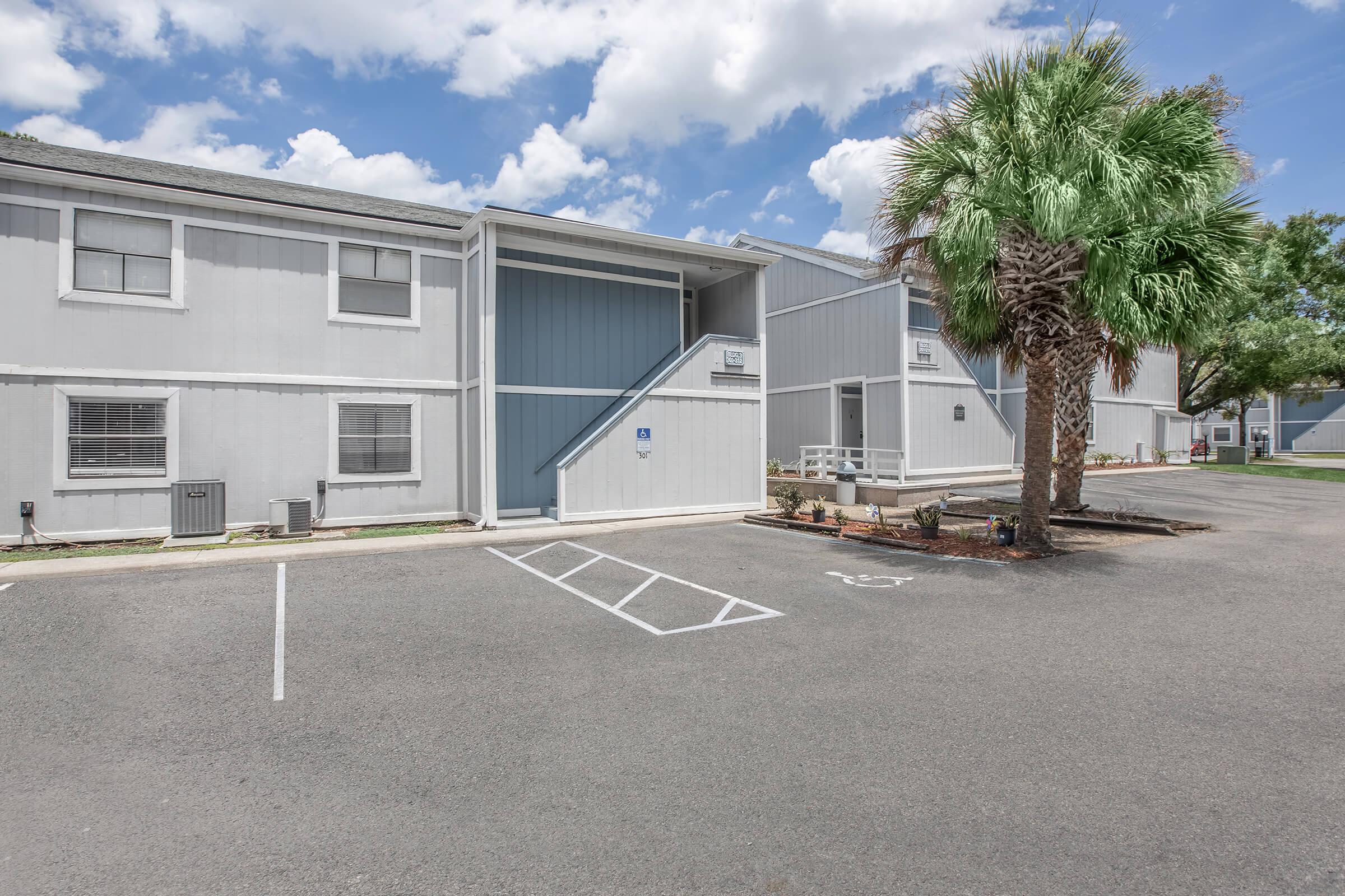 A residential building with a light gray exterior, featuring two separate units. Each unit has a set of stairs leading to the entrance, and there is a palm tree in the foreground. The parking lot has several marked spaces, and the sky is partly cloudy.