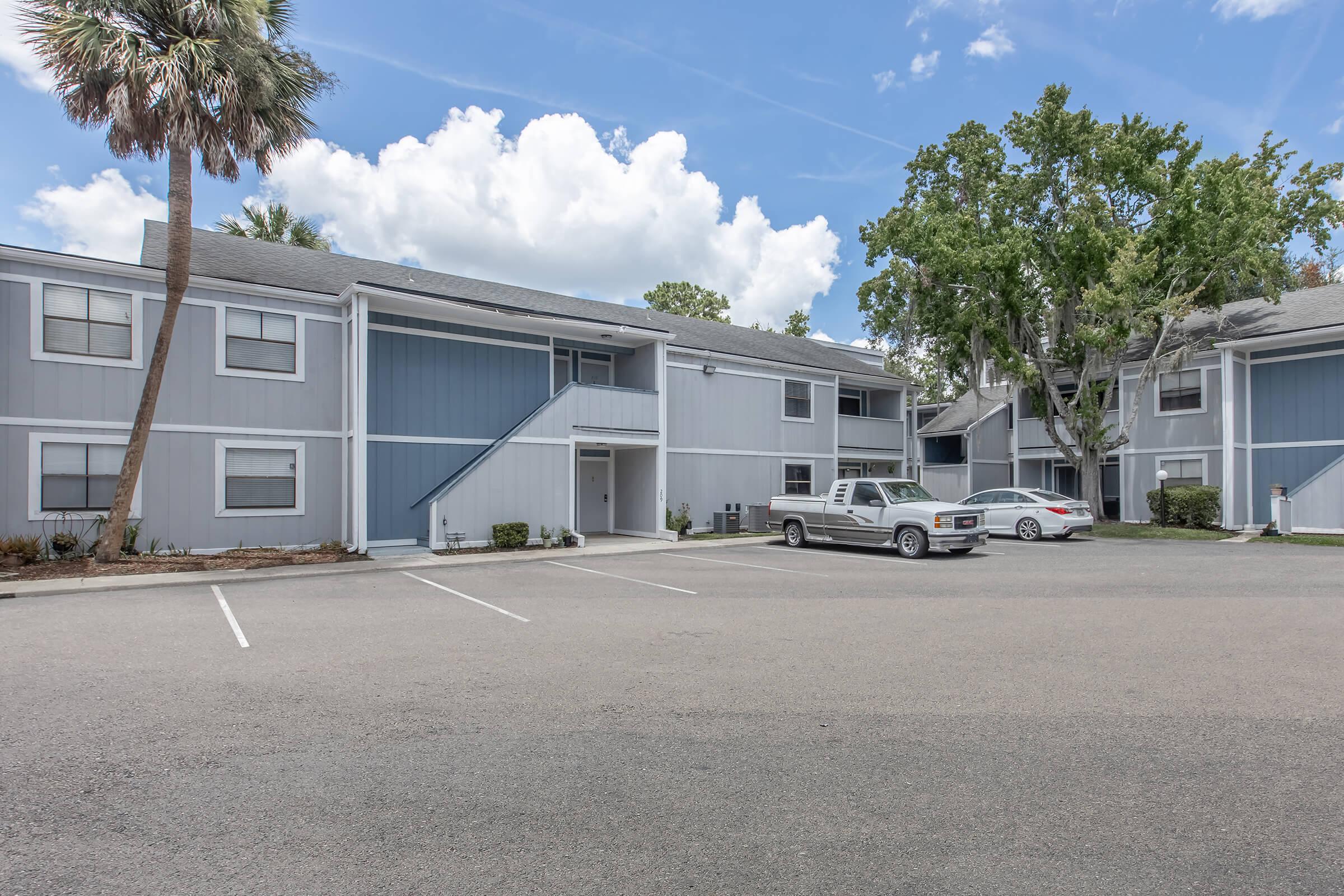A spacious parking lot in front of a two-story residential building with a light gray exterior. The building features multiple units, a staircase, and landscaping with palm trees. A few vehicles are parked nearby under a partly cloudy sky.