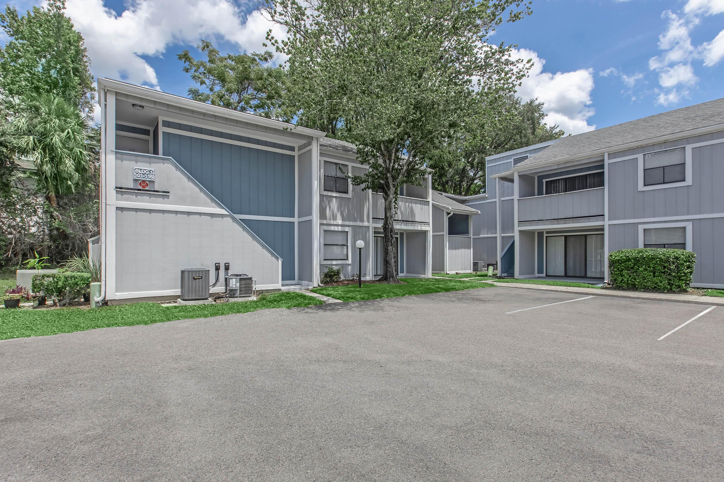 A view of a residential apartment complex featuring two-story buildings with gray and blue exteriors. The scene includes a grassy area, trees, and a parking lot with marked spaces. The sky is partly cloudy, adding to the pleasant, sunny ambiance of the area.