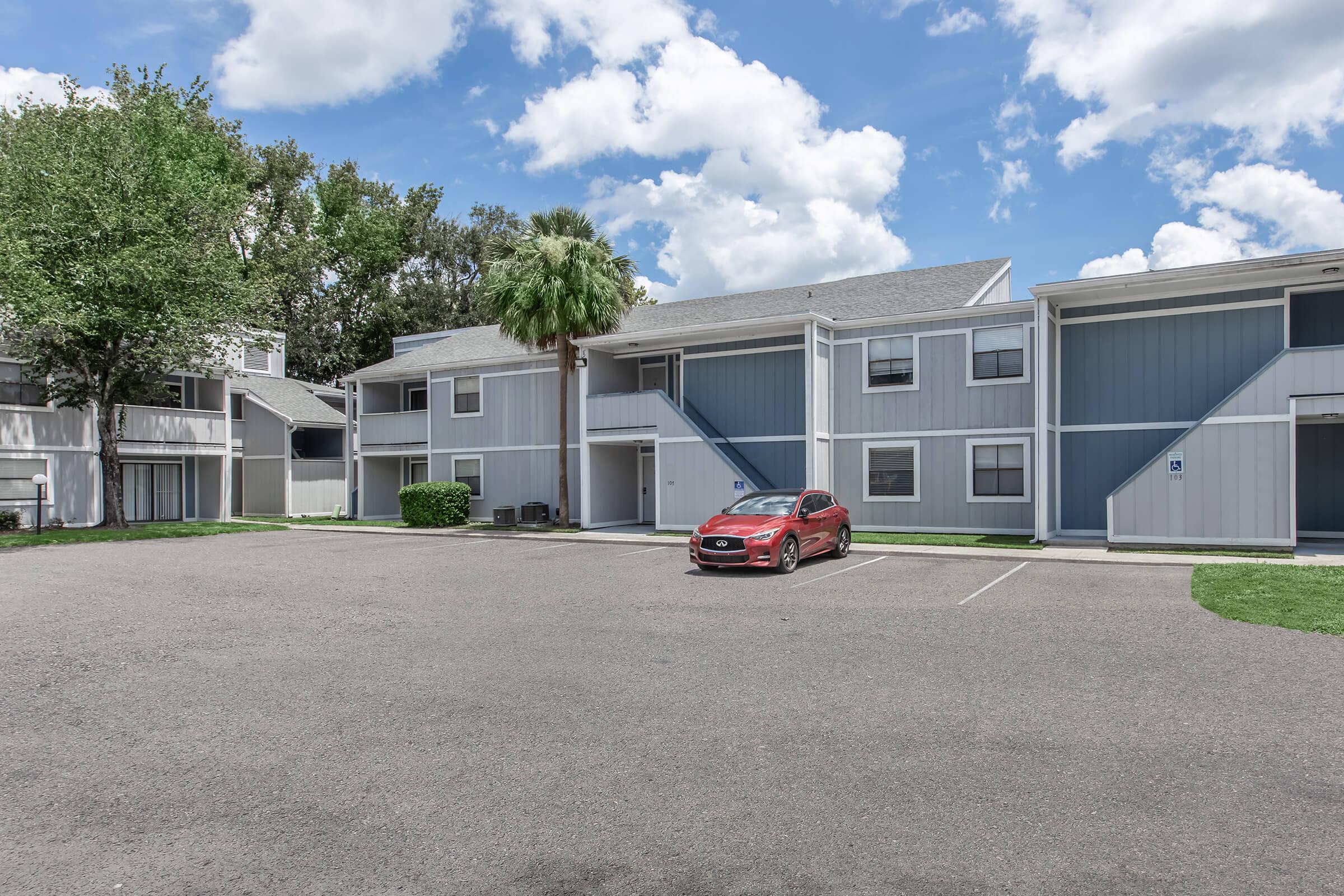 A view of a residential apartment complex with grey and blue exterior, featuring multiple two-story buildings. A red car is parked in the foreground on a gravel lot, surrounded by green grass and trees under a partly cloudy sky.