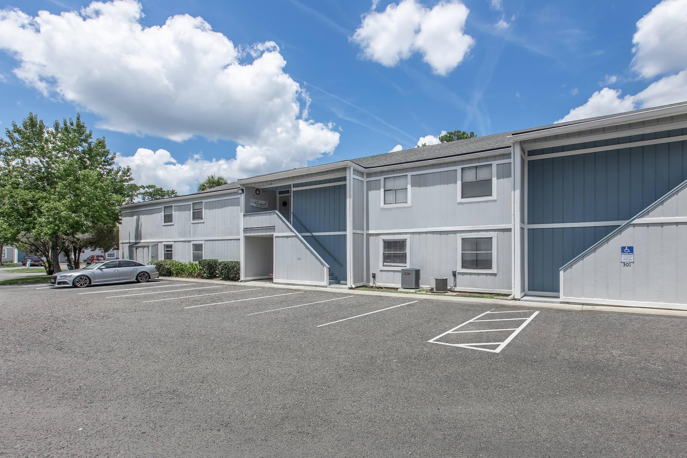 A view of a two-story apartment building with blue and white exteriors under a partly cloudy sky. Several parking spaces are visible in front, with a single car parked. Nearby, there are trees and landscaping, creating a welcoming atmosphere.