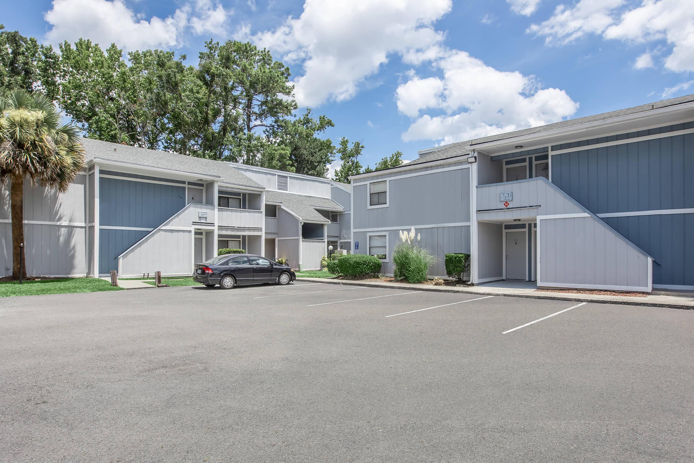 Two-story residential buildings with a modern design, featuring light gray and blue exteriors. The foreground shows a paved parking area with one parked black car. Lush green trees and shrubbery in the background, under a partly cloudy sky. 