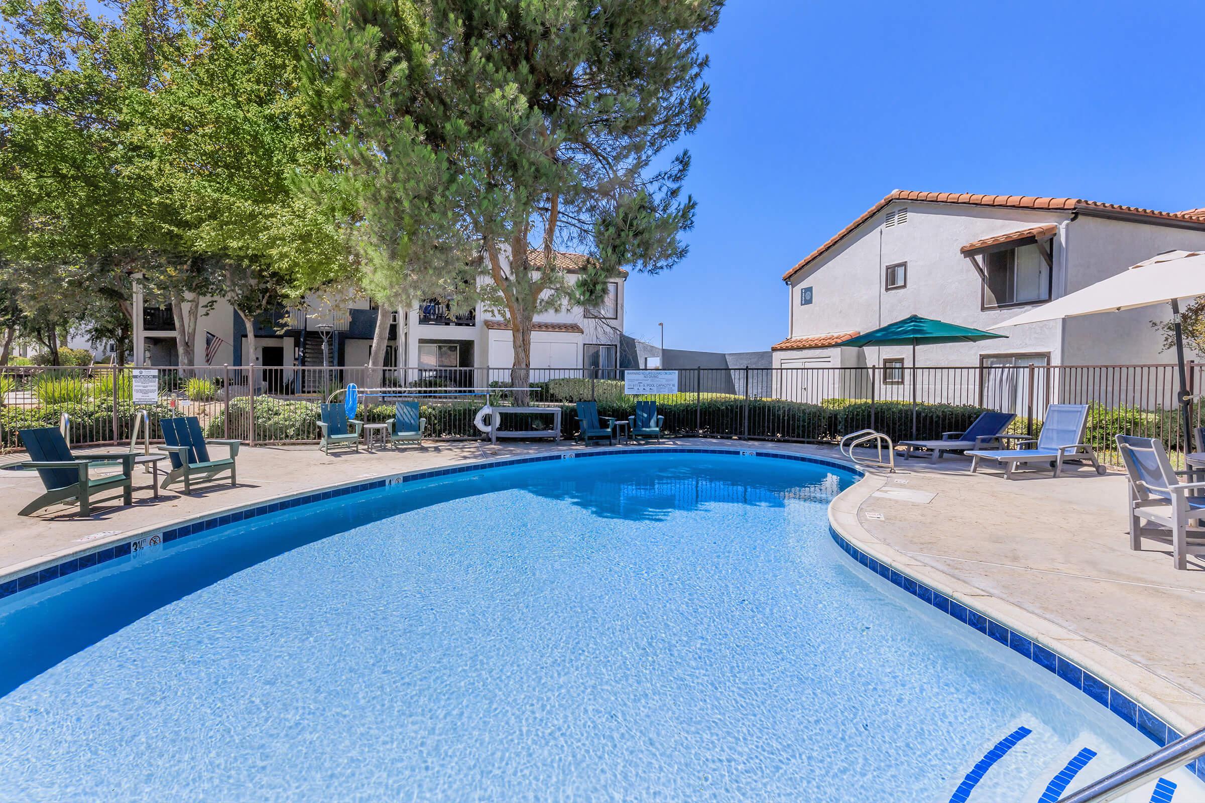 A clear blue swimming pool surrounded by lounge chairs and umbrellas, with lush greenery in the background. Nearby buildings feature a light-colored exterior and a tiled roof, contributing to a relaxing outdoor atmosphere. The sky is bright and clear.
