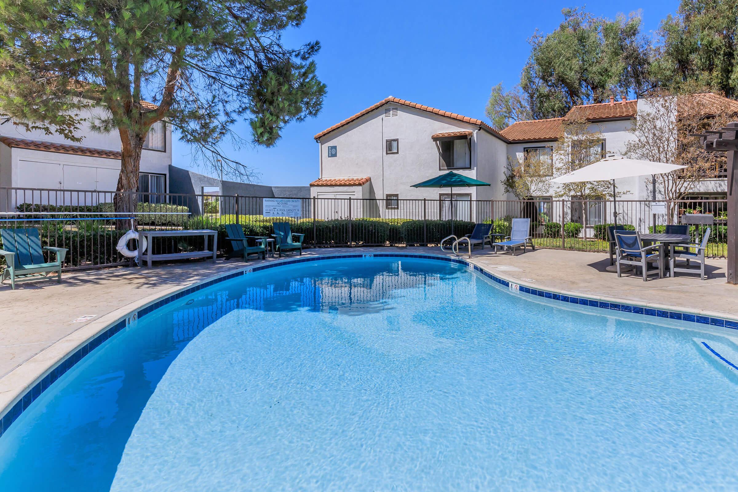 A sparkling blue swimming pool surrounded by lounge chairs and umbrellas, with a backdrop of two-story residential buildings and lush greenery under a clear blue sky. The scene conveys a relaxing outdoor atmosphere perfect for leisure.