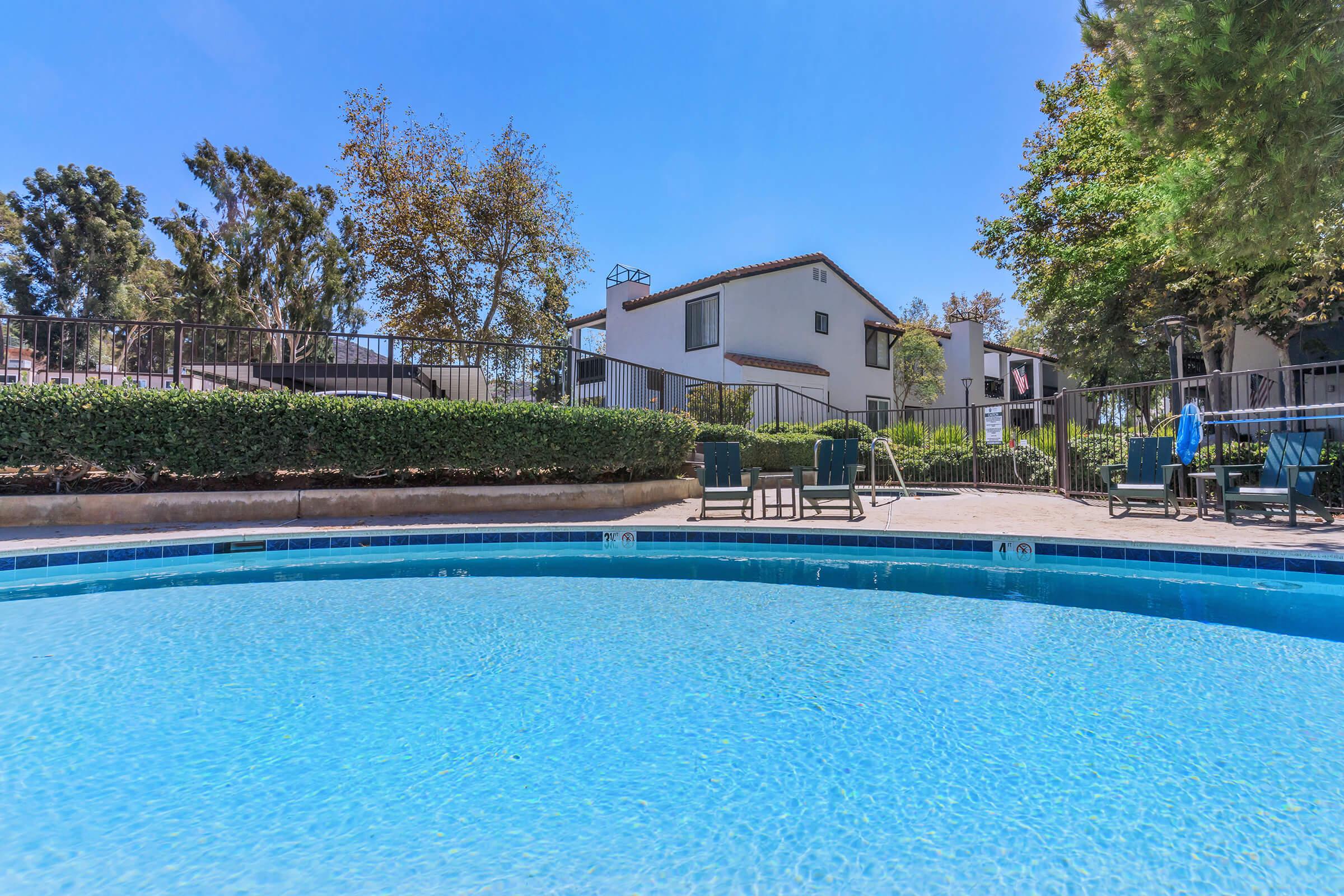 A clear blue swimming pool with a circular edge, surrounded by lush greenery and a fence. In the background, there are white buildings and trees under a bright blue sky. Several lounge chairs are positioned near the pool, creating a relaxing outdoor atmosphere.