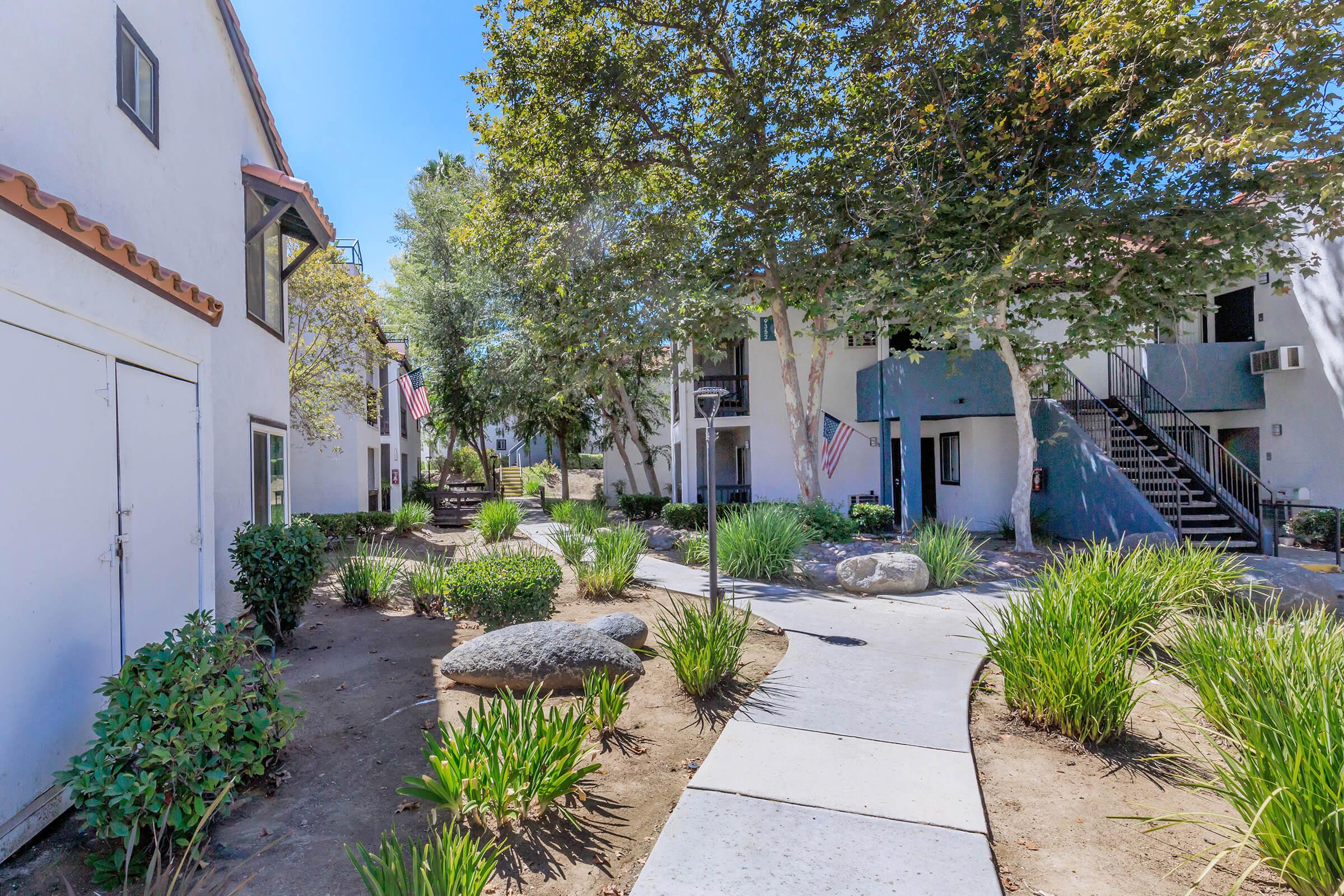 A walkway lined with green shrubs and grasses, leading through a residential area with white buildings. The scene is well-lit by sunlight, with trees providing shade and a few decorative rocks placed along the path. In the background, there are stairs leading to upper floors of the buildings.