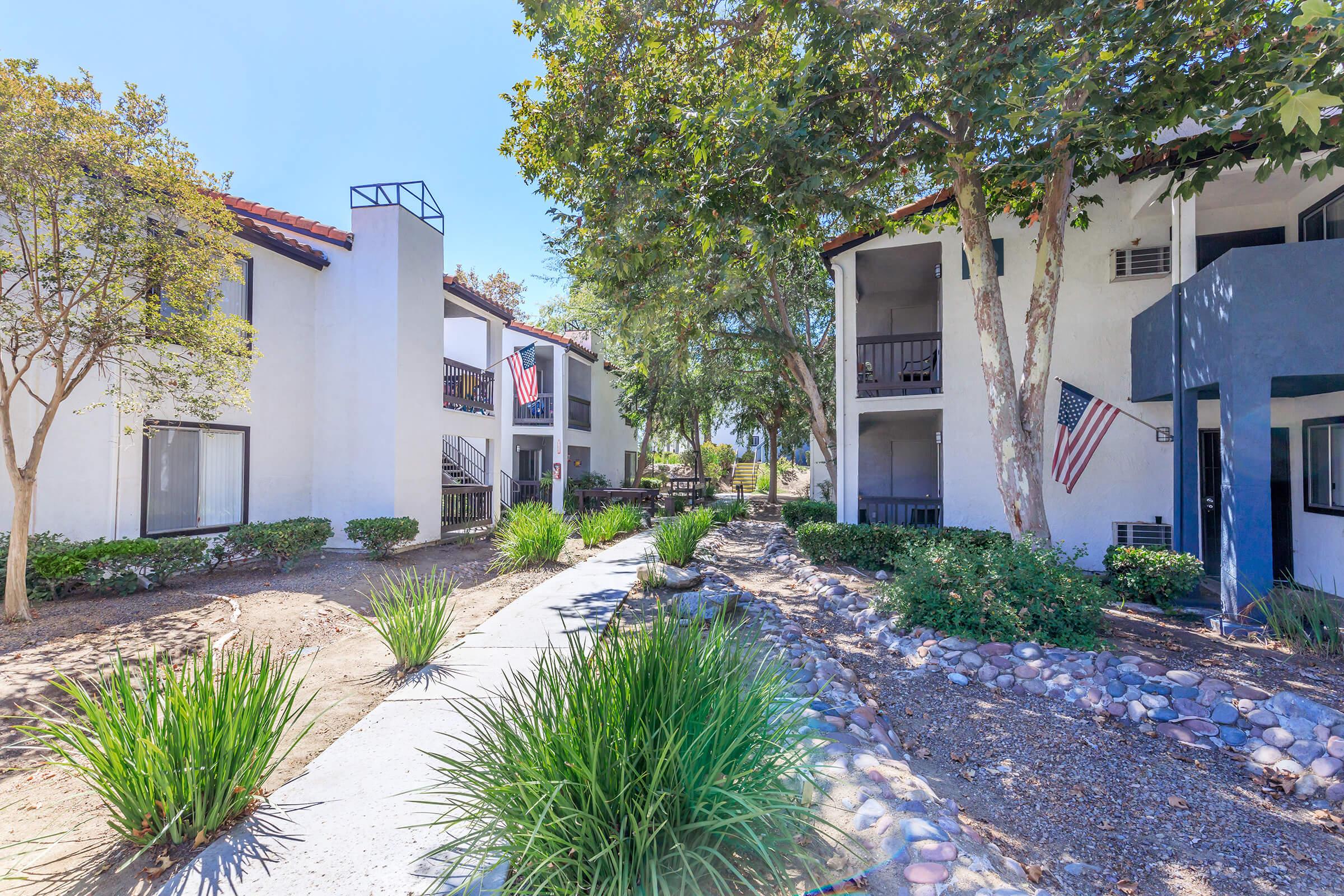 A well-maintained apartment complex surrounded by greenery, featuring two buildings with balconies. A clear walkway lined with plants leads through the complex, showcasing American flags on some balconies against a bright blue sky.