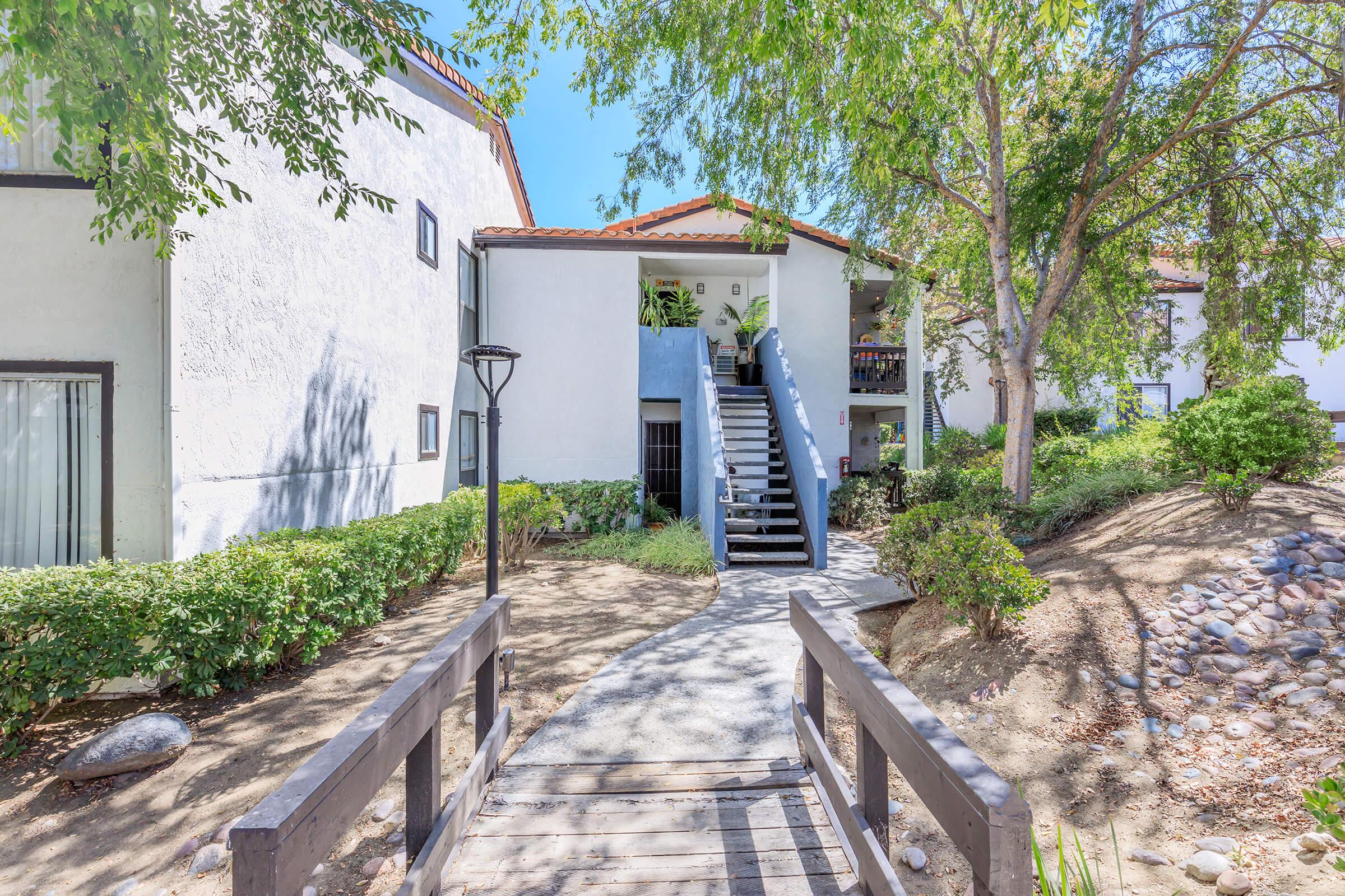 A well-maintained apartment complex with a staircase leading to the upper level, surrounded by green bushes and trees. A wooden bridge crosses over a small pathway, leading to the entrance. Bright blue sky visible above, creating a welcoming atmosphere.