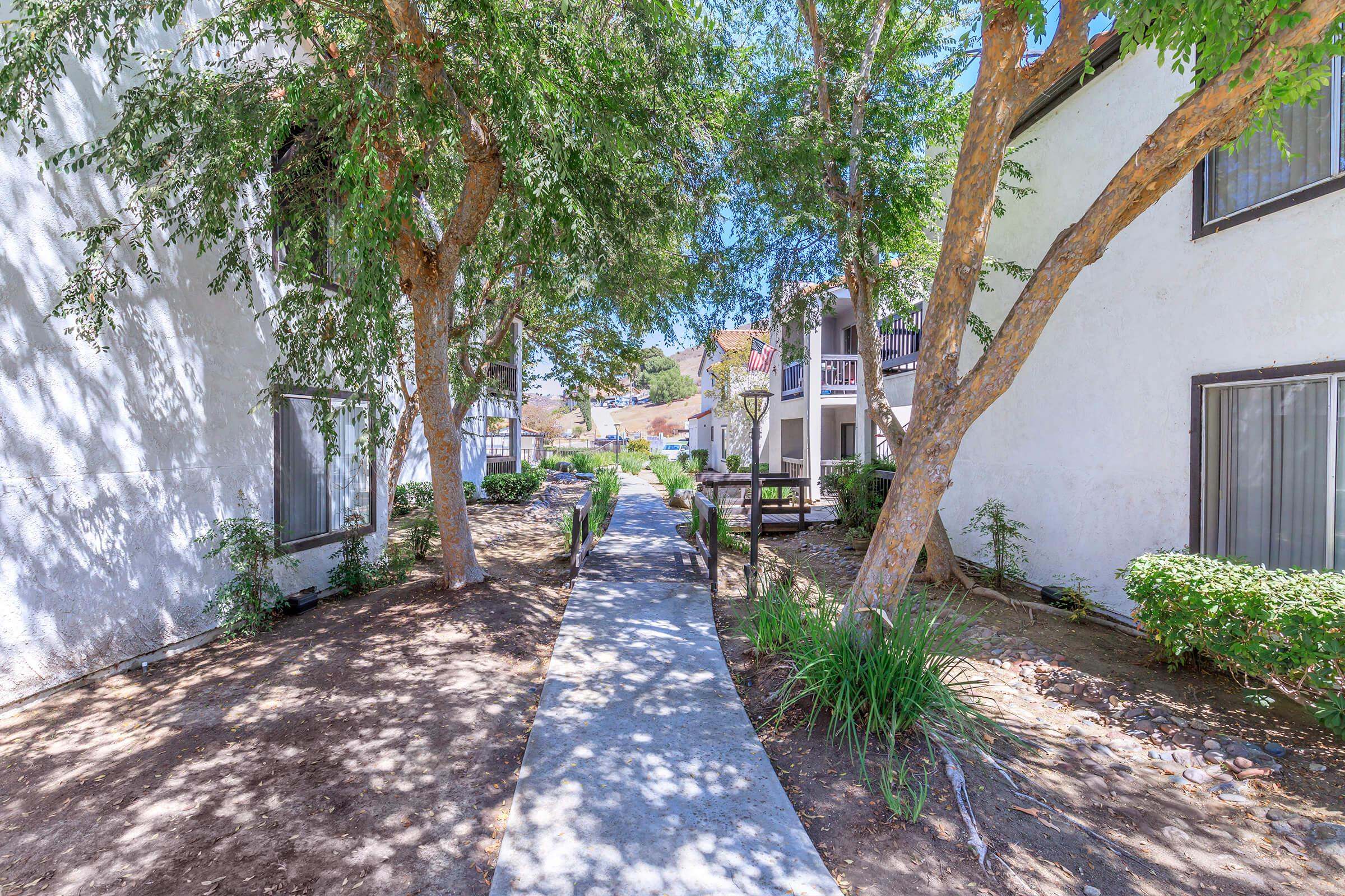 A concrete pathway lined with trees leads through a landscaped area between two white apartment buildings. Lush green plants and shrubs are visible, creating a serene outdoor atmosphere. The scene is bright and inviting, with clear blue skies above. The pathway appears to lead further into the complex.