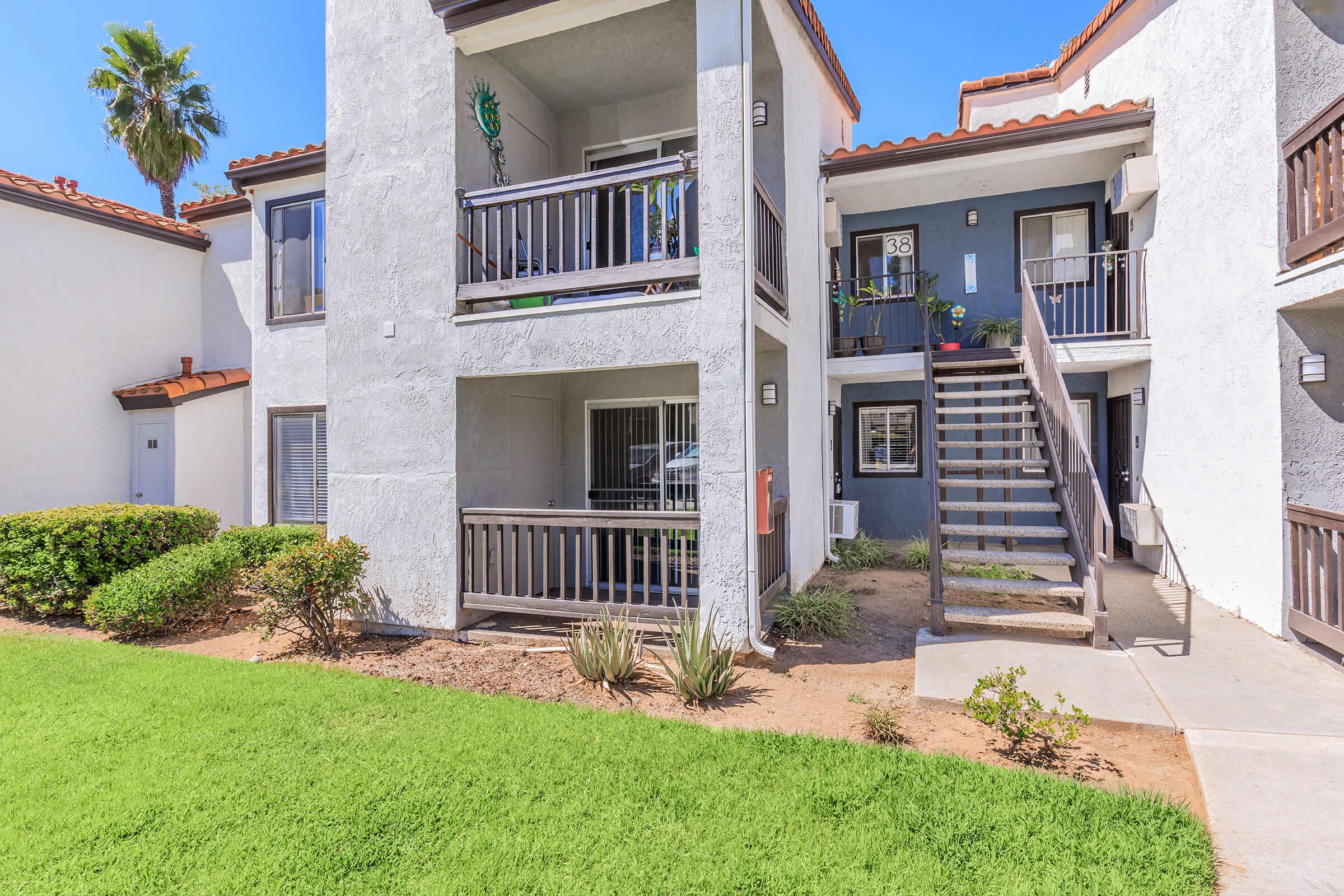 Two-story apartment building with a concrete exterior and balconies. The ground level has a staircase leading to the upper apartments. There is a small landscaped area with grass and shrubs in front. Bright blue sky and sunshine indicate a clear day.
