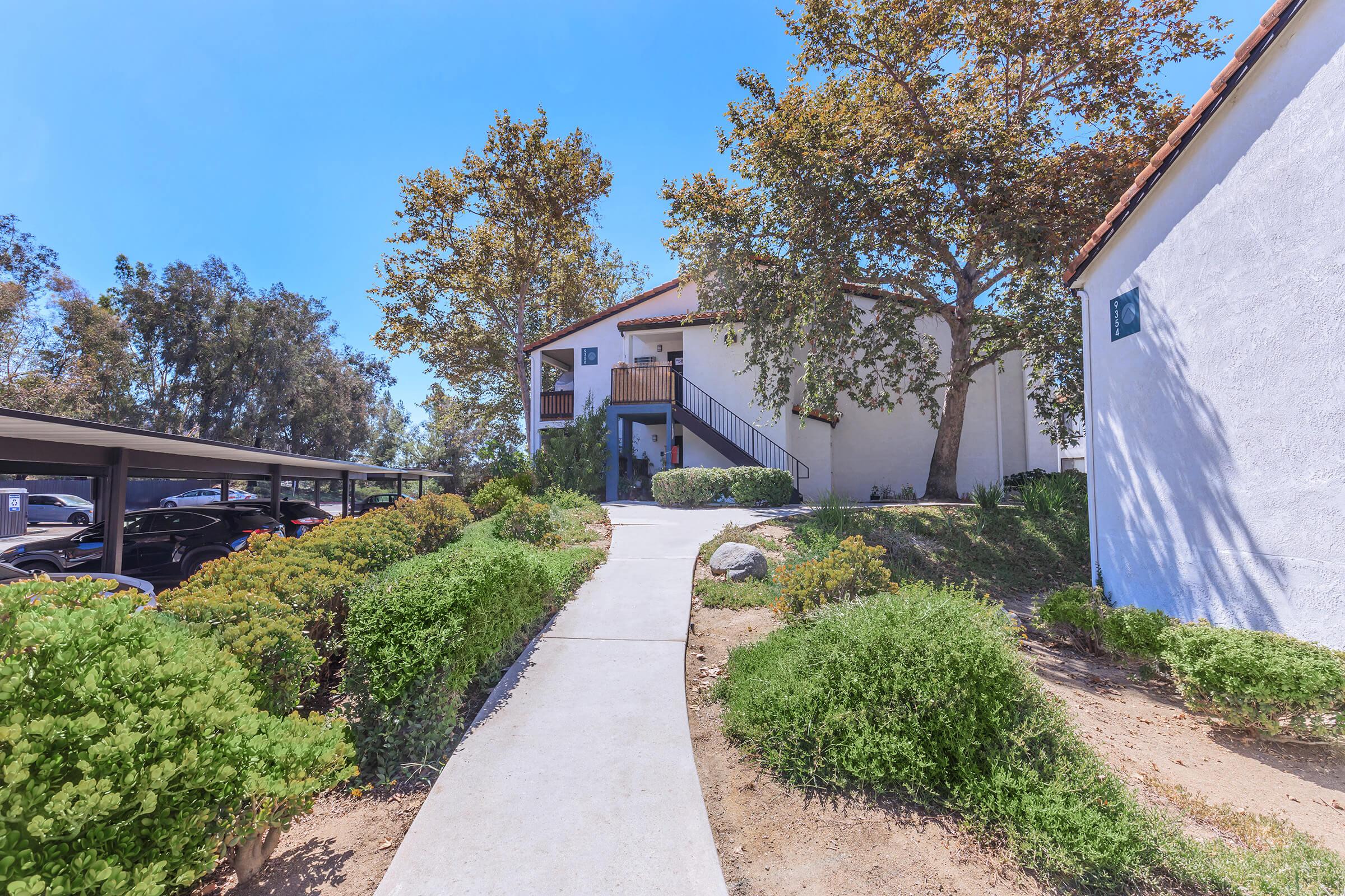 A walkway leading to a white multi-unit building surrounded by lush greenery and trees. There are neatly trimmed shrubs lining the path, and parking spaces are visible on the left under a carport. The sky is clear and blue, indicating a sunny day.