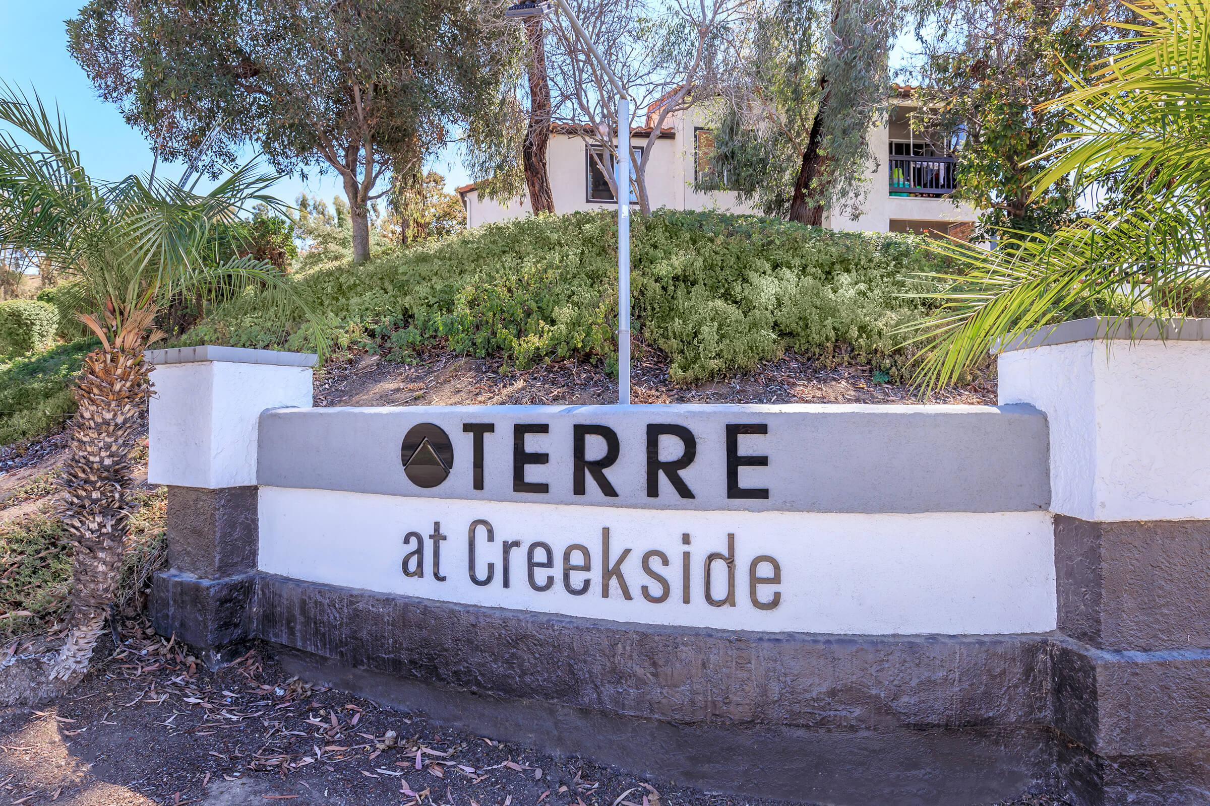 A sign displaying "TERRE at Creekside," situated in a landscaped area with greenery and palm trees. The sign features a modern design with bold lettering, indicating a residential or commercial property named Creekside. 