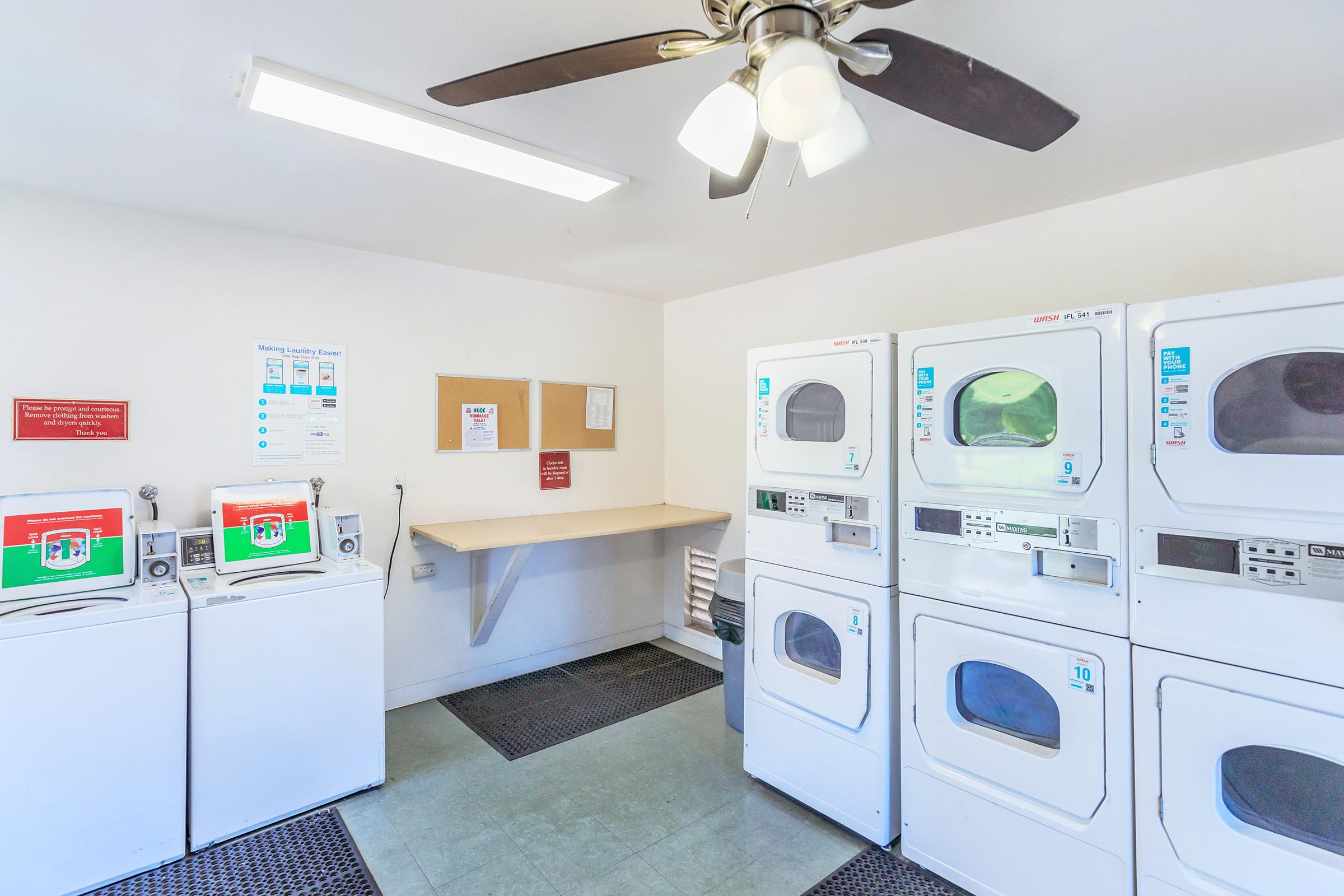 Interior of a laundry room featuring several white washing machines and dryers stacked against the wall. There is a ceiling fan, a small table for folding clothes, bulletin boards with information, and a few mats on the floor. The space is well-lit and organized for laundry use.