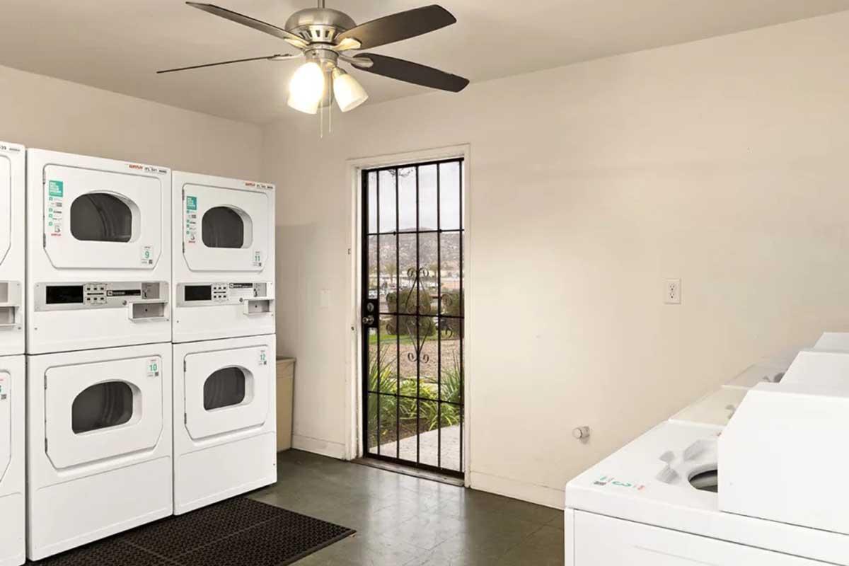 A laundry room featuring multiple stacked washer and dryer units, a ceiling fan with lights, and a door with metal security bars leading outside. The space has a simple design with neutral colors and a matted entryway.