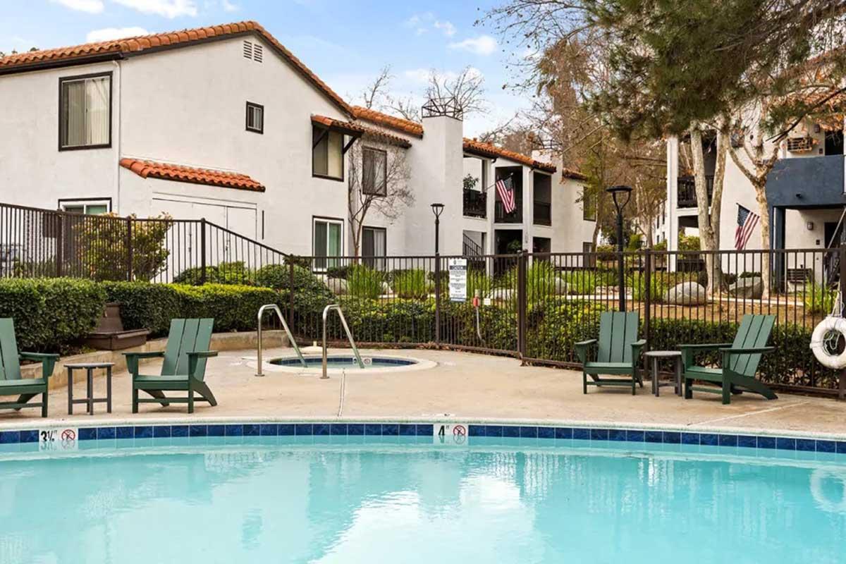 A sunny outdoor pool area surrounded by lush greenery, featuring poolside lounge chairs and a hot tub. The backdrop includes residential buildings with a Mediterranean architectural style and American flags. The area is enclosed with a fence, creating a relaxing atmosphere.