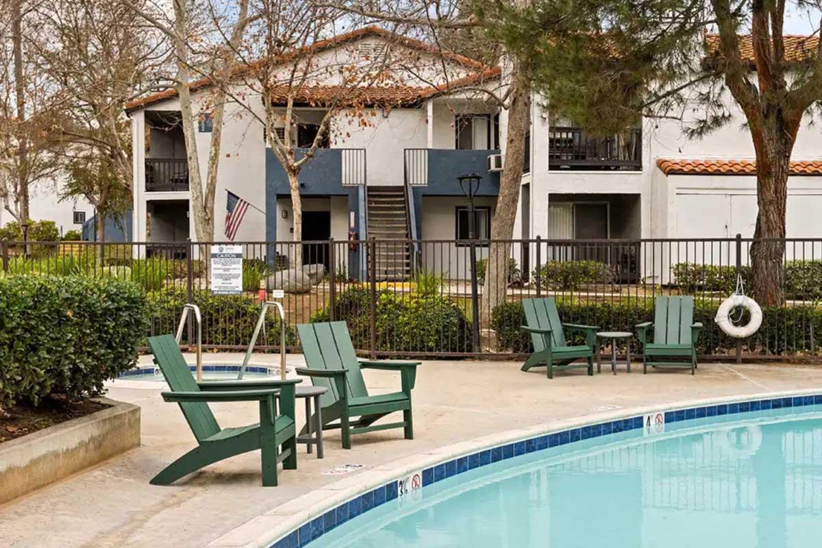 A swimming pool area featuring green Adirondack chairs beside the water. In the background, there are two-story apartments with balconies and an American flag. Surrounding the pool are hedges and trees, creating a relaxing outdoor setting.