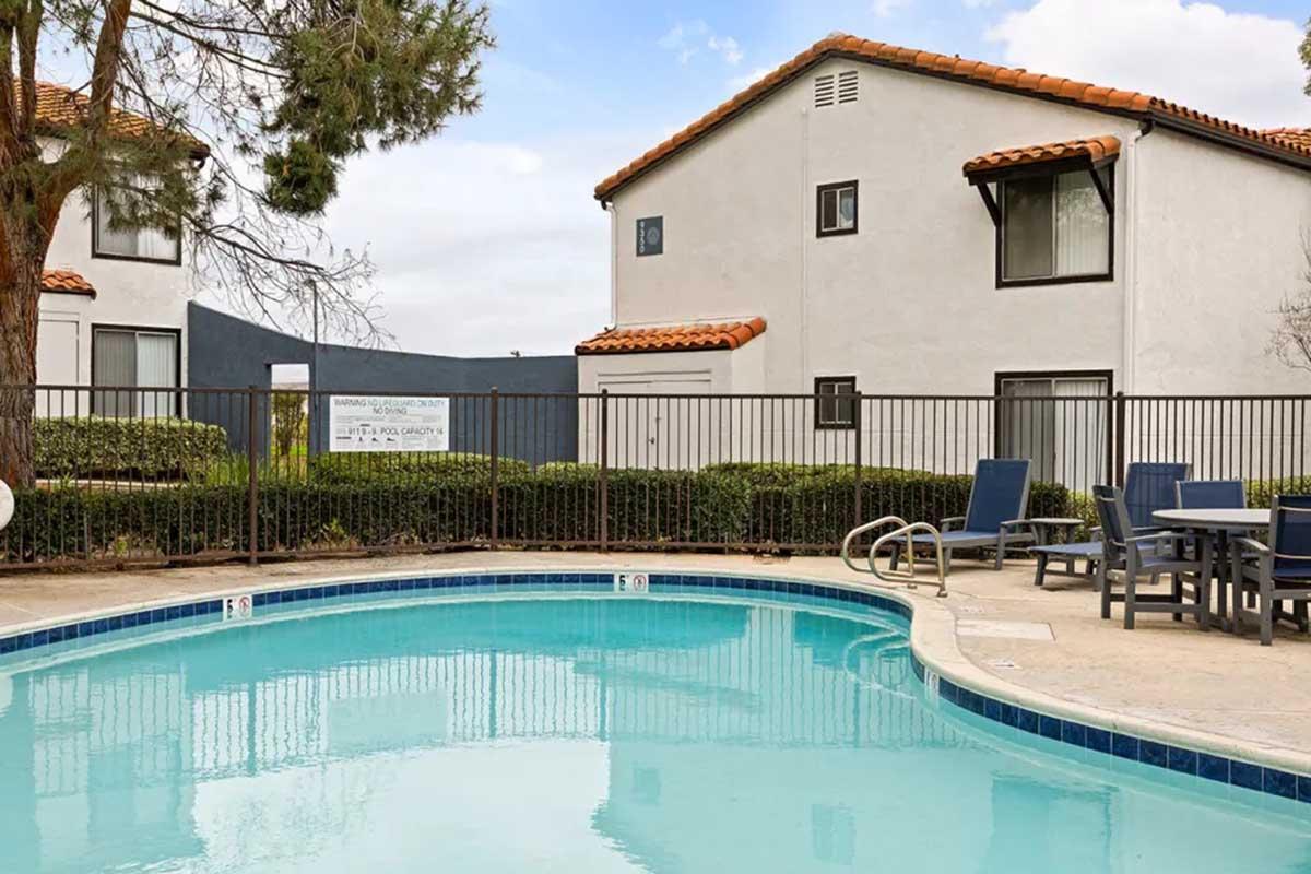 A swimming pool surrounded by a fence, with lounge chairs and tables nearby. In the background, there are two white buildings with red-tiled roofs and greenery. The sky is partly cloudy, creating a serene atmosphere.